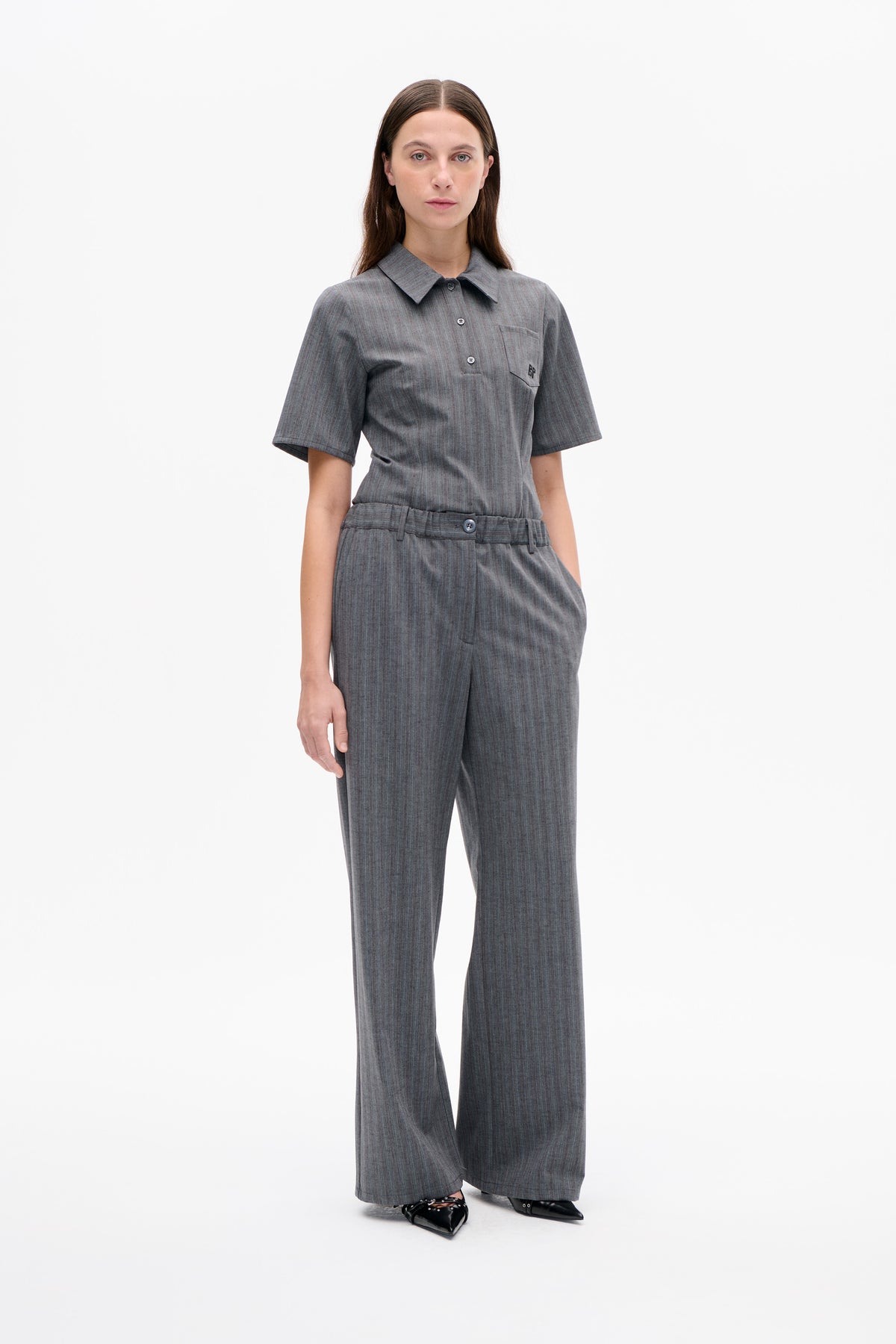 Frontalansicht eines Models, das ein graues, nadelgestreiftes Kurzarmhemd mit Knopfleiste und Brusttasche sowie eine passende, weit geschnittene Hose trägt.