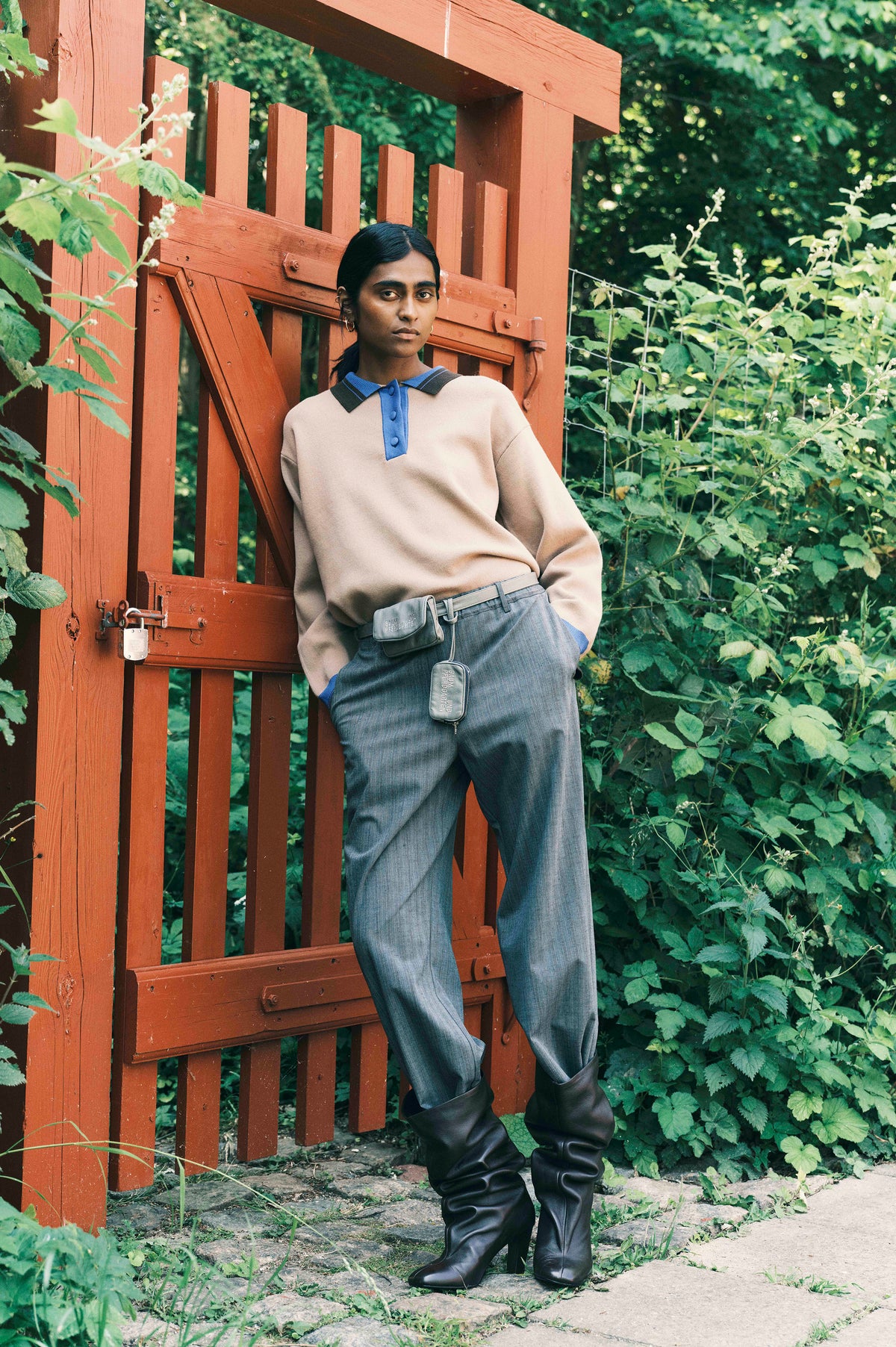 Kampagnenporträt eines Models in einem cremefarbenen Pullover mit blau-anthrazitfarbenem Kragen, einer grauen Nadelstreifenhose und schwarzen Stiefeln, das an einem roten Holztor lehnt und von Grün umgeben ist.