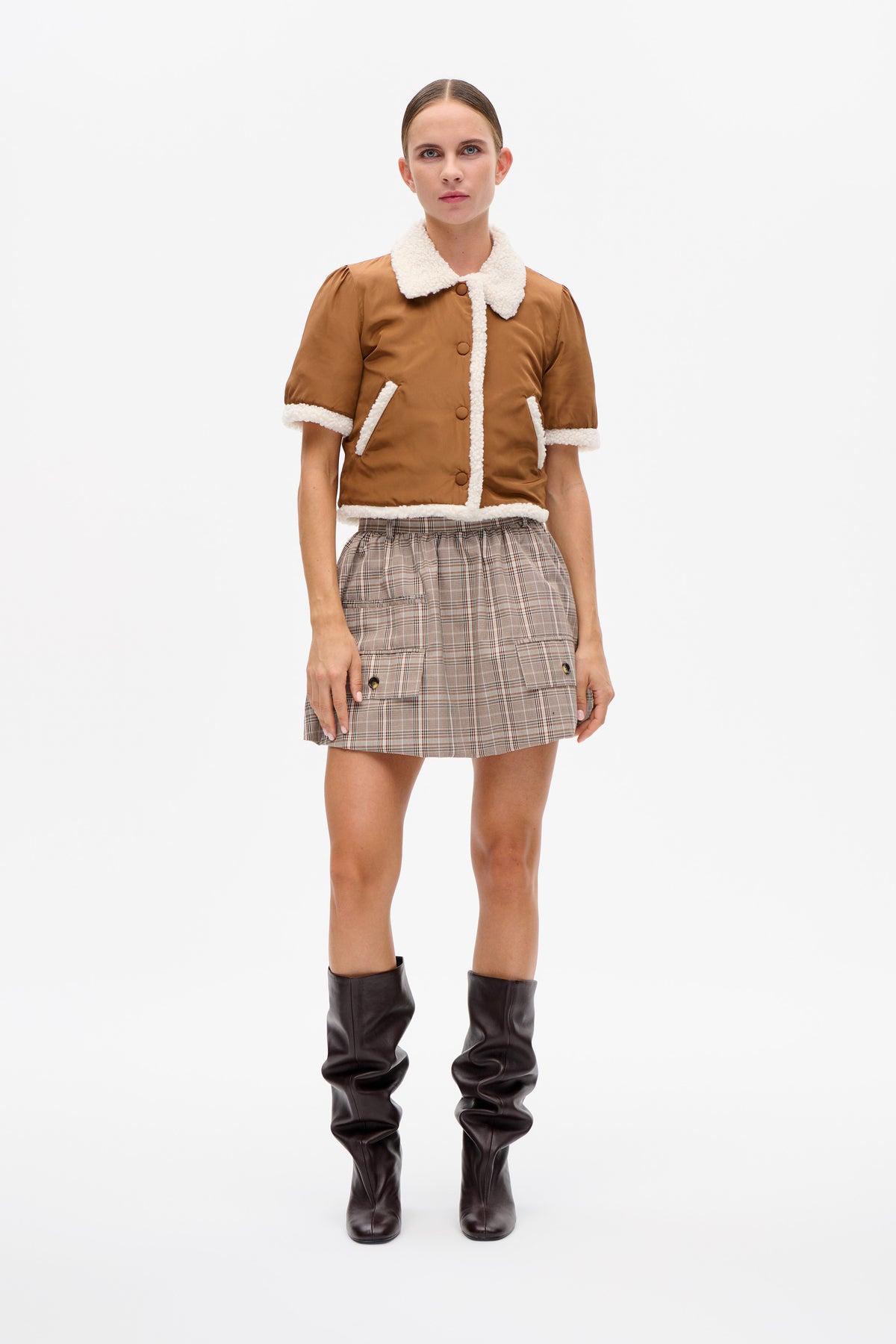Model draagt een karamelbruin jasje met korte mouwen en een pluizige crèmekleurige afwerking, een beige geruite minirok en donkerkleurige, losvallende kniehoge laarzen.