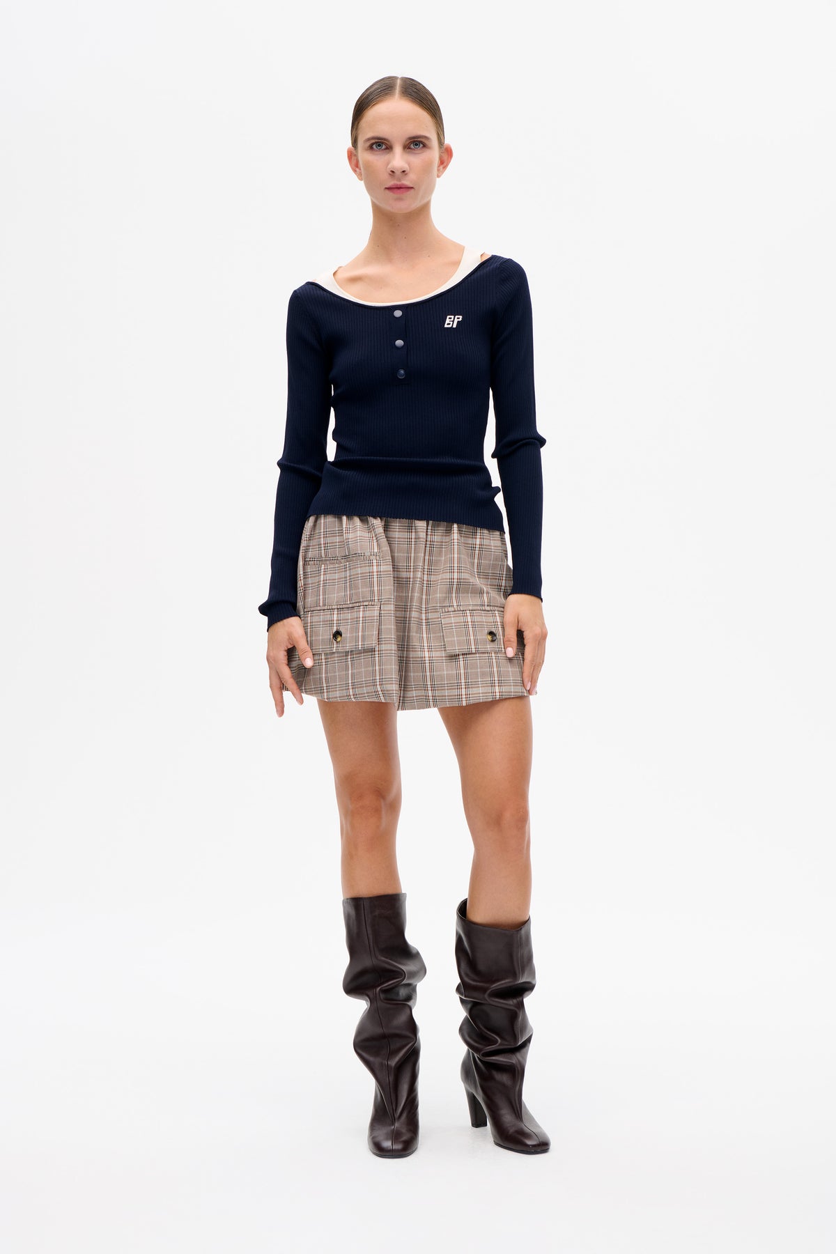 Model wearing a navy ribbed button-front top with white neckline trim, beige plaid shorts and slouchy dark brown knee-high boots.