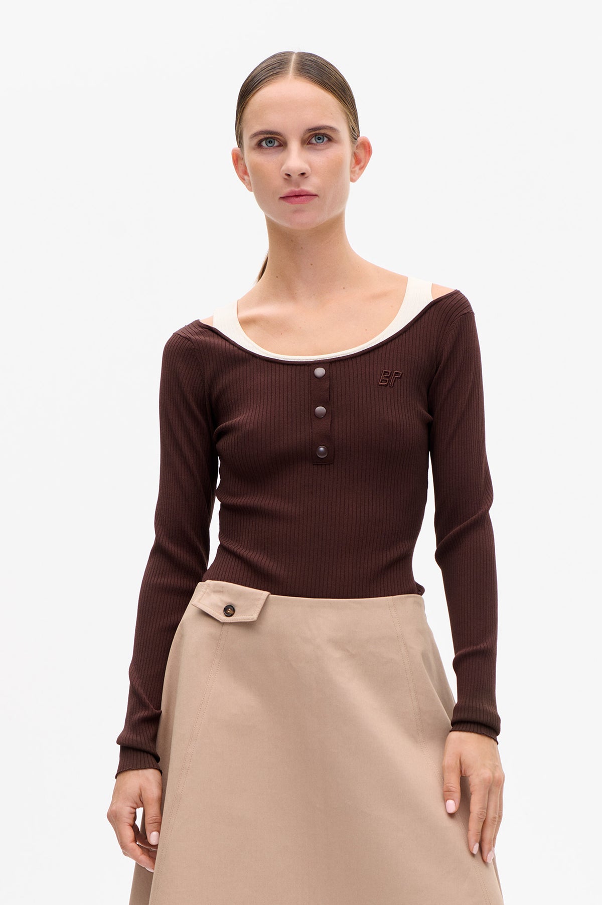 Front view of a model wearing a dark brown ribbed long-sleeve top with a wide neckline, snap buttons and a small chest emblem over a cream top.