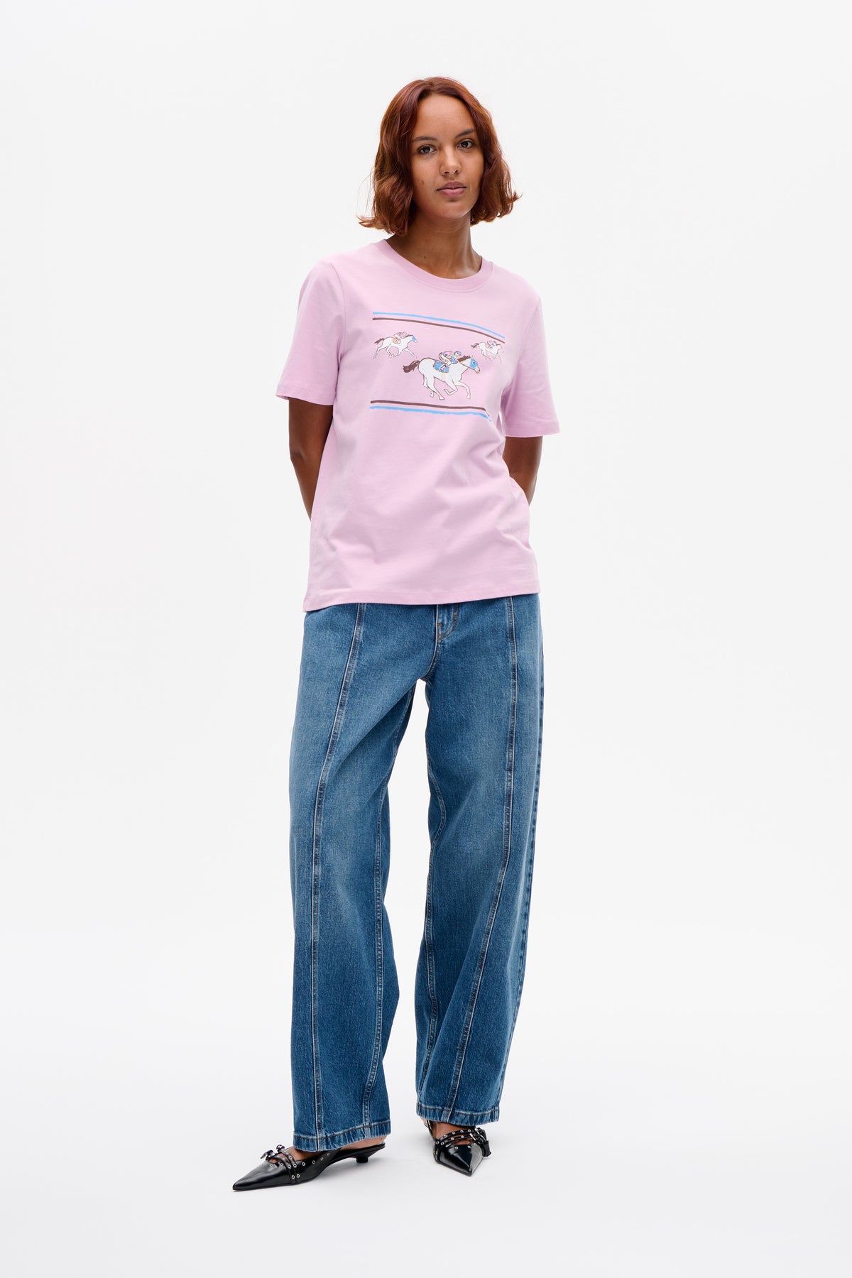 Model wearing a pale pink short-sleeve T-shirt with a graphic of galloping horses and blue and brown stripes, paired with blue jeans.