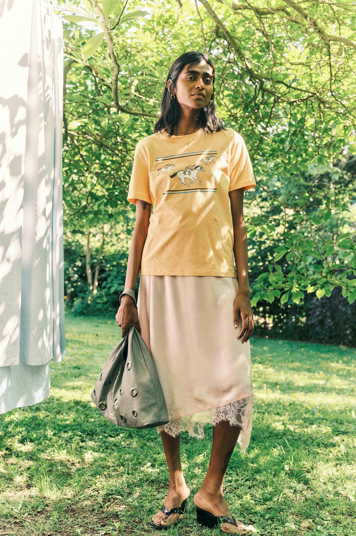 Model outdoors wearing a pale yellow T-shirt with horse illustrations and a pale skirt with a lace hem, carrying a grey eyelet bag.