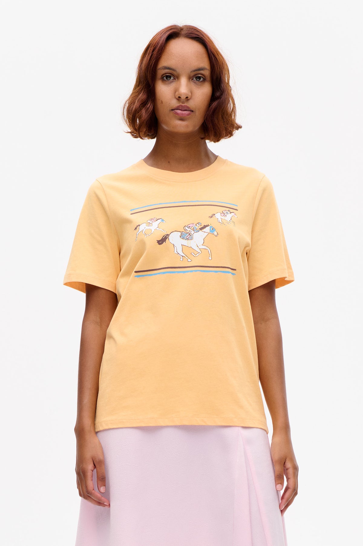 Front view of a pale yellow short-sleeved T-shirt with a crew neckline and colorful horse-racing graphic, paired with a pale pink skirt.