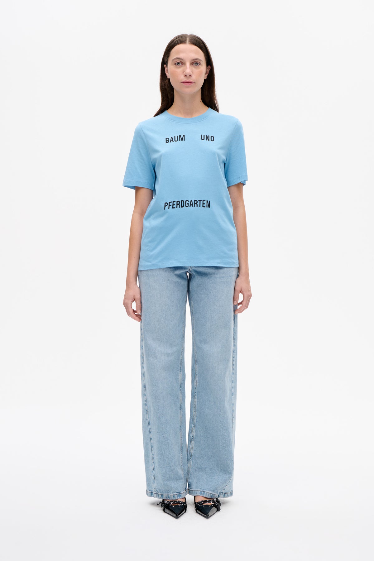 Modèle portant un T-shirt bleu clair à manches courtes avec lettrage noir Baum und Pferdgarten, associé à un jean délavé clair à jambes larges.