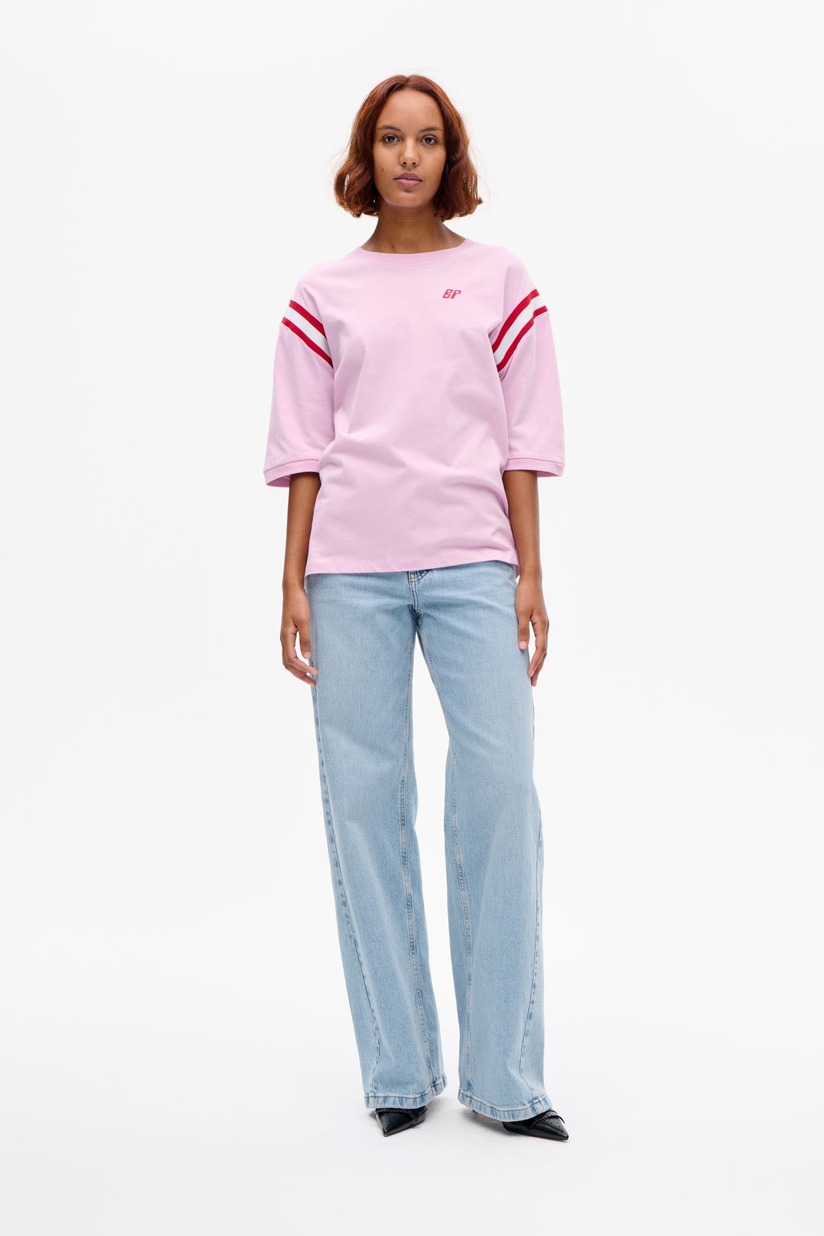 Vorderansicht eines Models, das ein hellrosa kurzärmeliges T-Shirt mit roten Ärmelstreifen und kleinem Brustlogo trägt, kombiniert mit hellblauen Jeans.