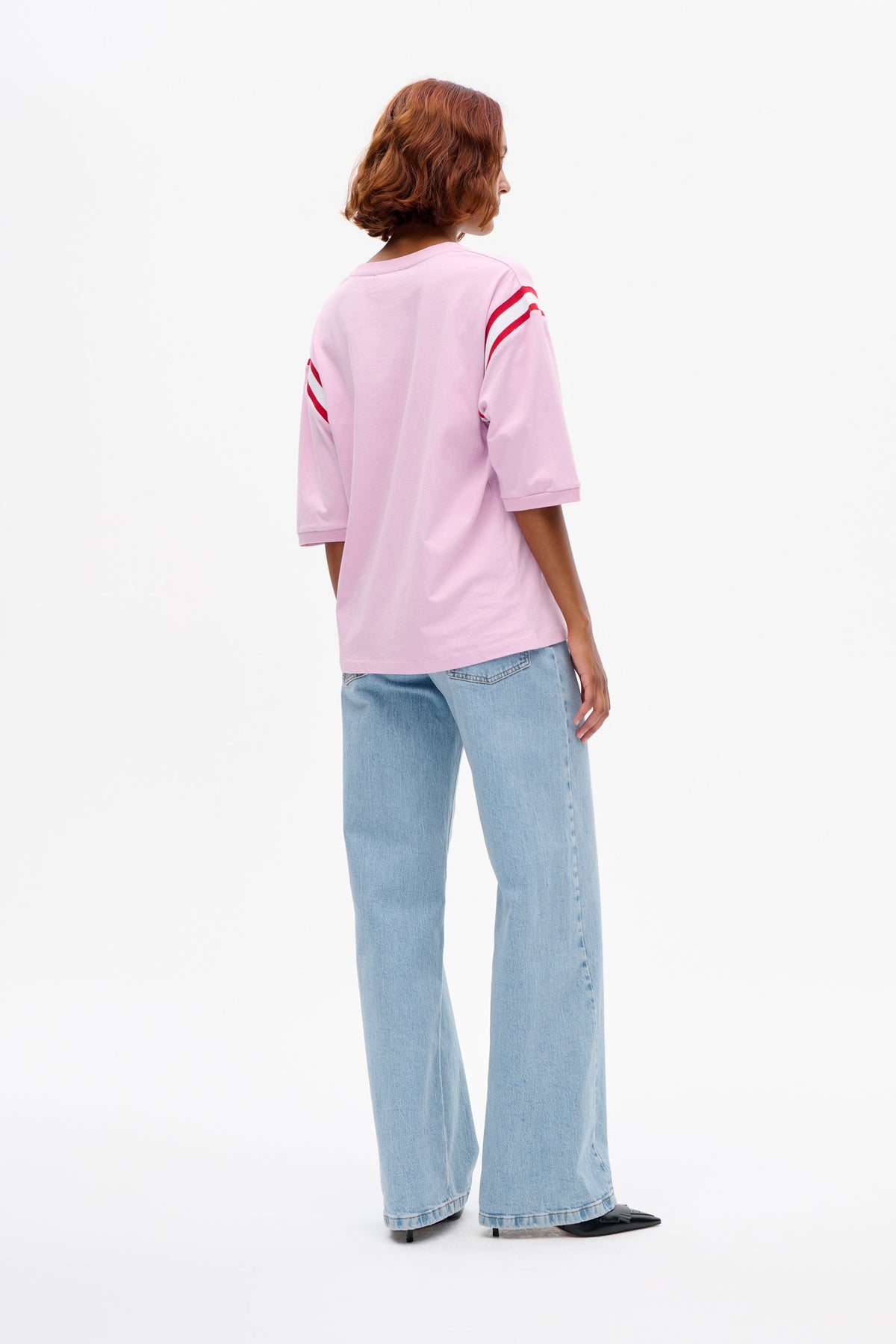 Rückenansicht eines Models, das ein blassrosa T-Shirt mit roten und weißen Streifen an den Ärmeln sowie eine weit geschnittene Jeans in heller Waschung trägt.