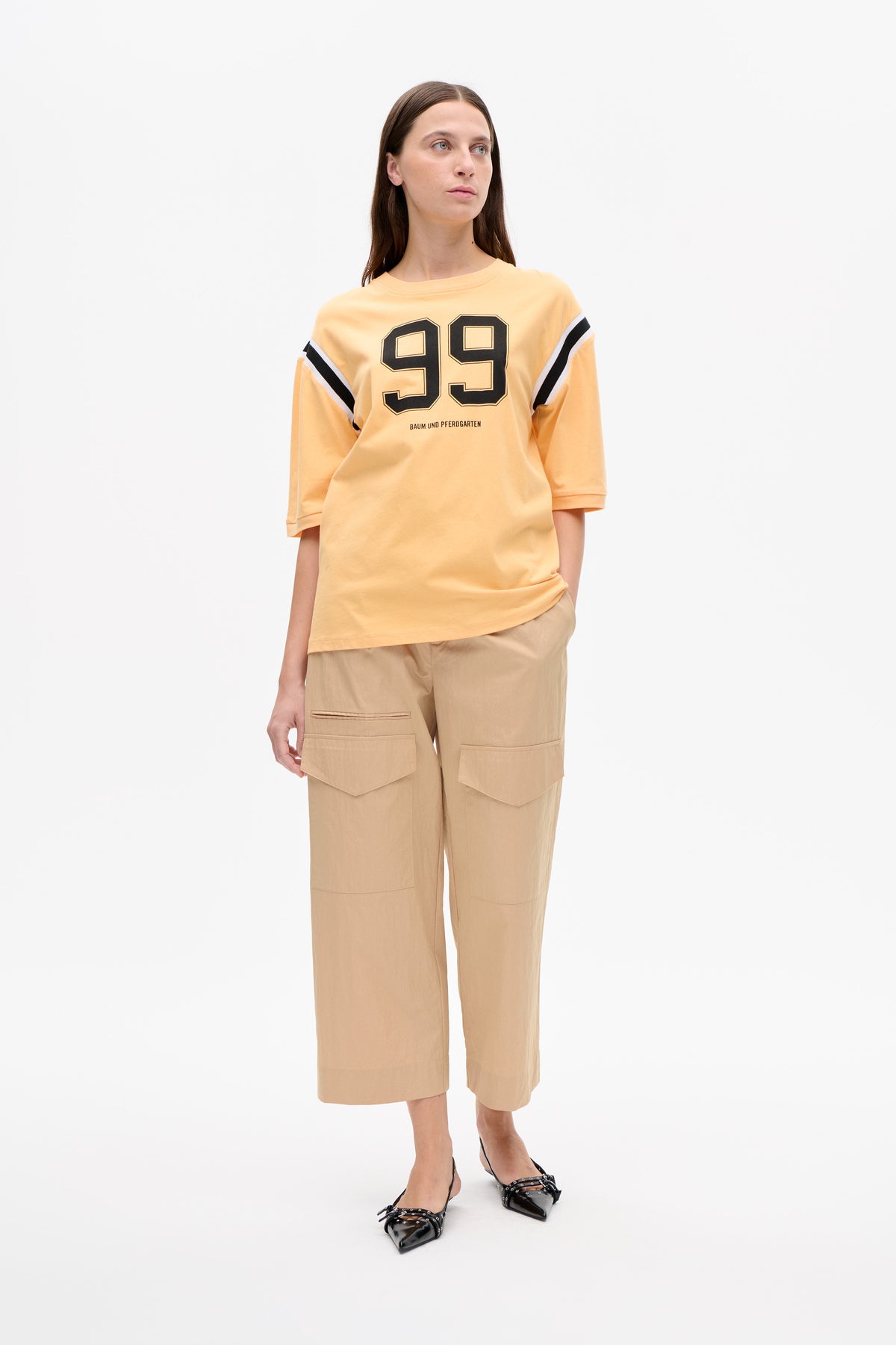 Model wearing a peach oversized short-sleeve top with a black 99 graphic and striped sleeves, paired with beige wide-leg cropped trousers.