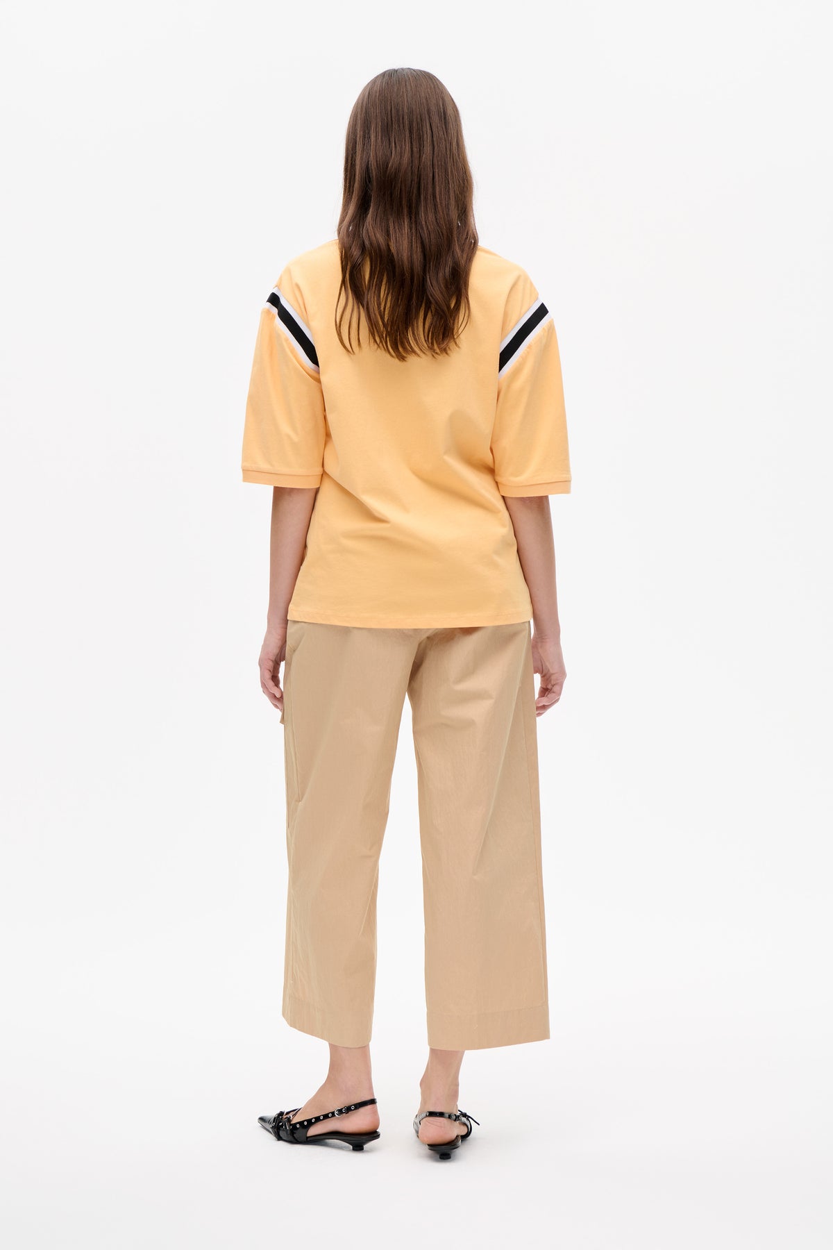 Back view of a model wearing a pale yellow short-sleeve top with black and white shoulder stripes and beige cropped wide-leg trousers.