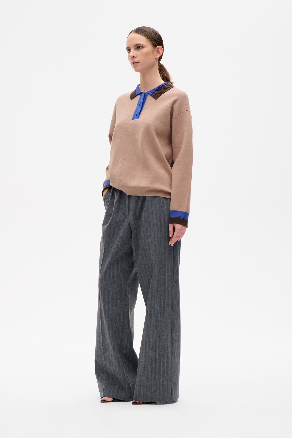 Model wearing a beige long-sleeved knit polo with blue trim and a brown striped collar, paired with charcoal pinstripe wide-leg trousers.