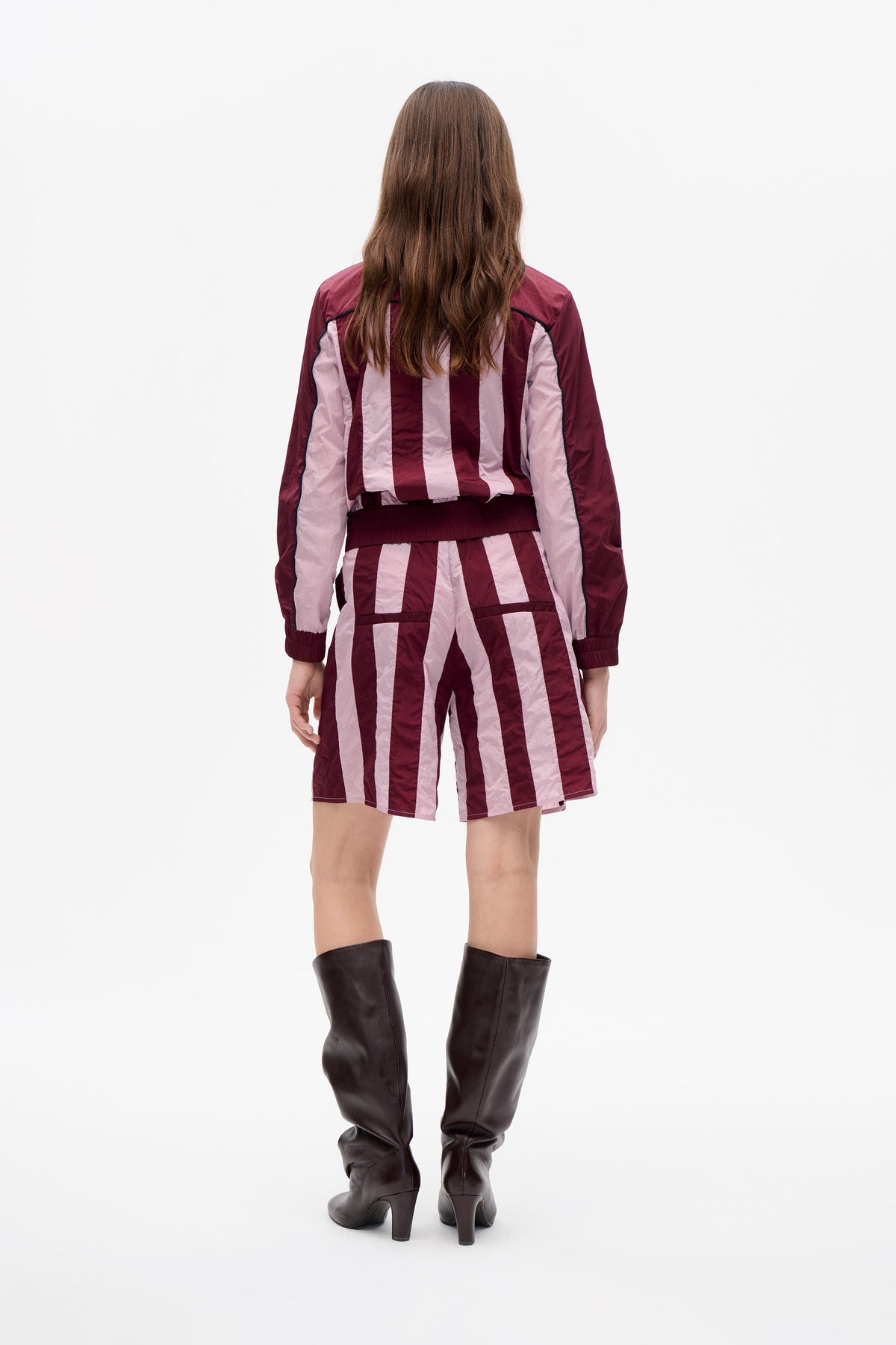 Back view of a model in a burgundy and pale pink striped jacket and matching shorts, with dark piping and tall brown boots.