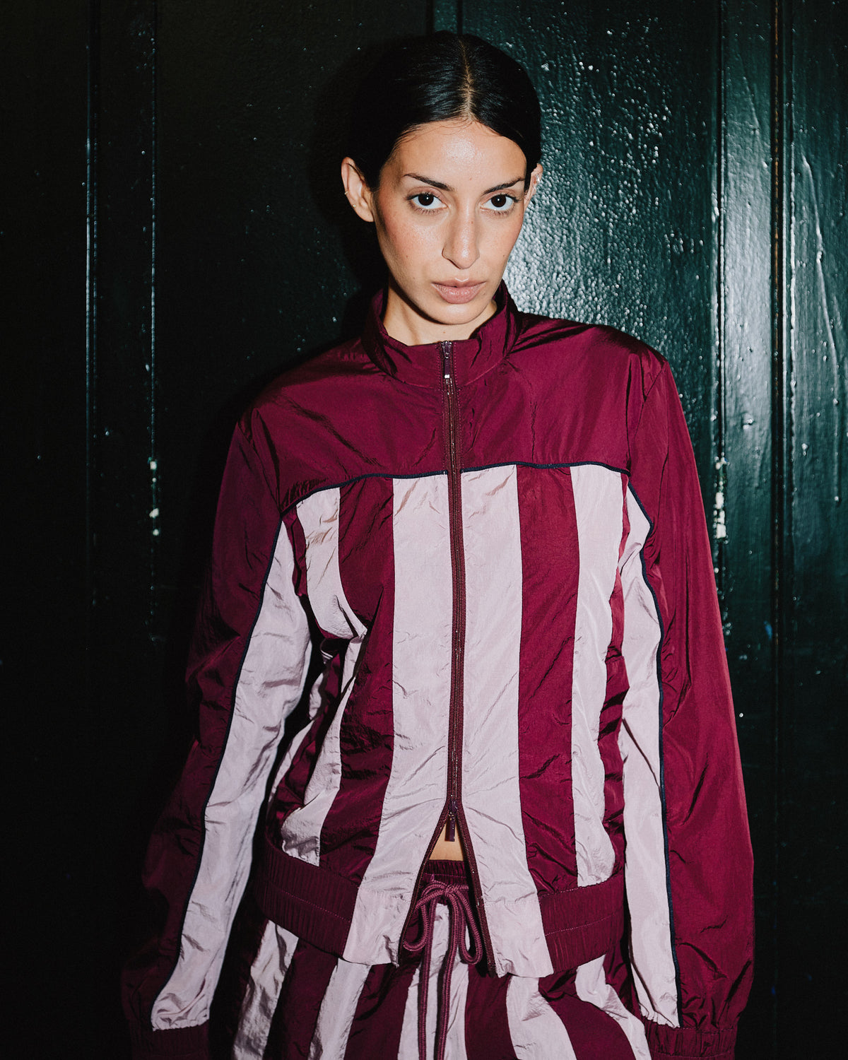 Campaign portrait of a model wearing a burgundy zip-front track jacket with broad pale pink vertical panels and matching trousers against a dark, wet wall.