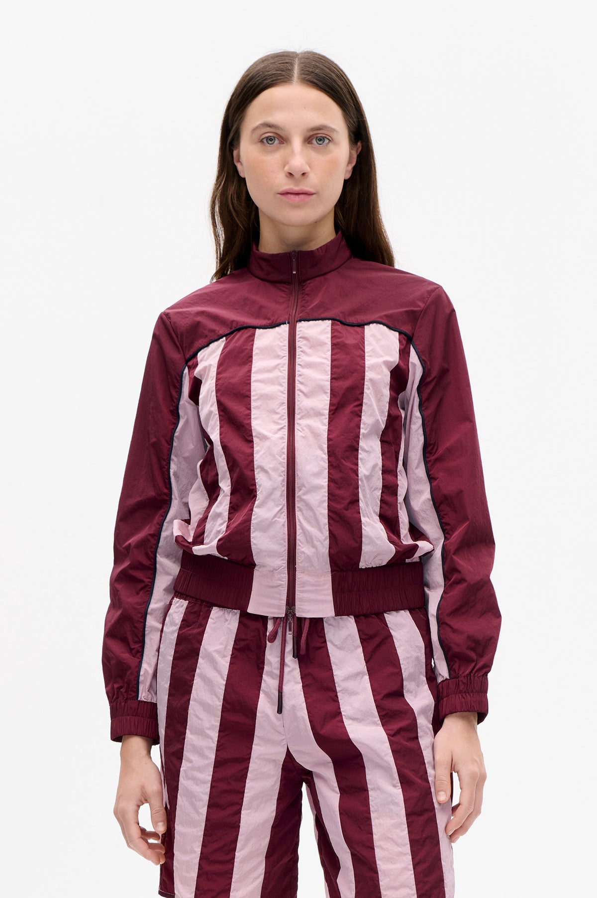 Front view of a burgundy zip-up jacket with a high collar, pale pink vertical stripes, dark piping and elastic cuffs.