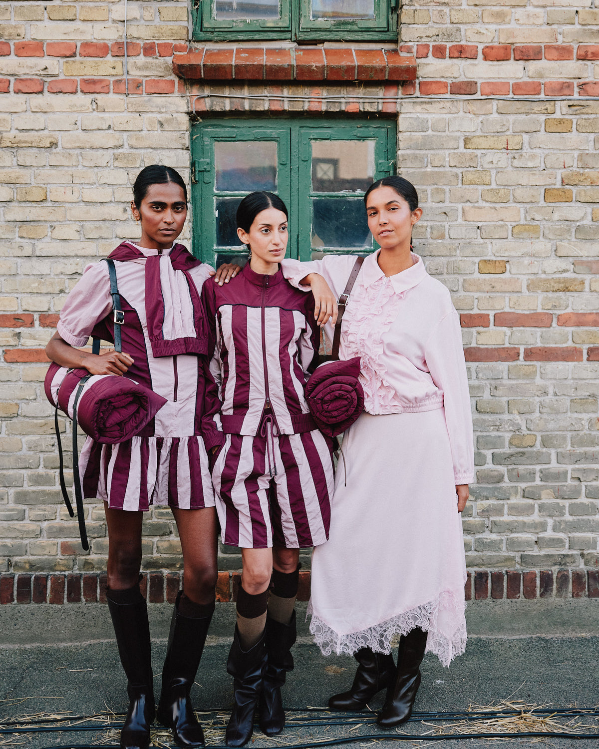 Three models pose against a brick wall in burgundy-and-pale-pink striped outfits and a pale pink ruffled dress with a lace hem.