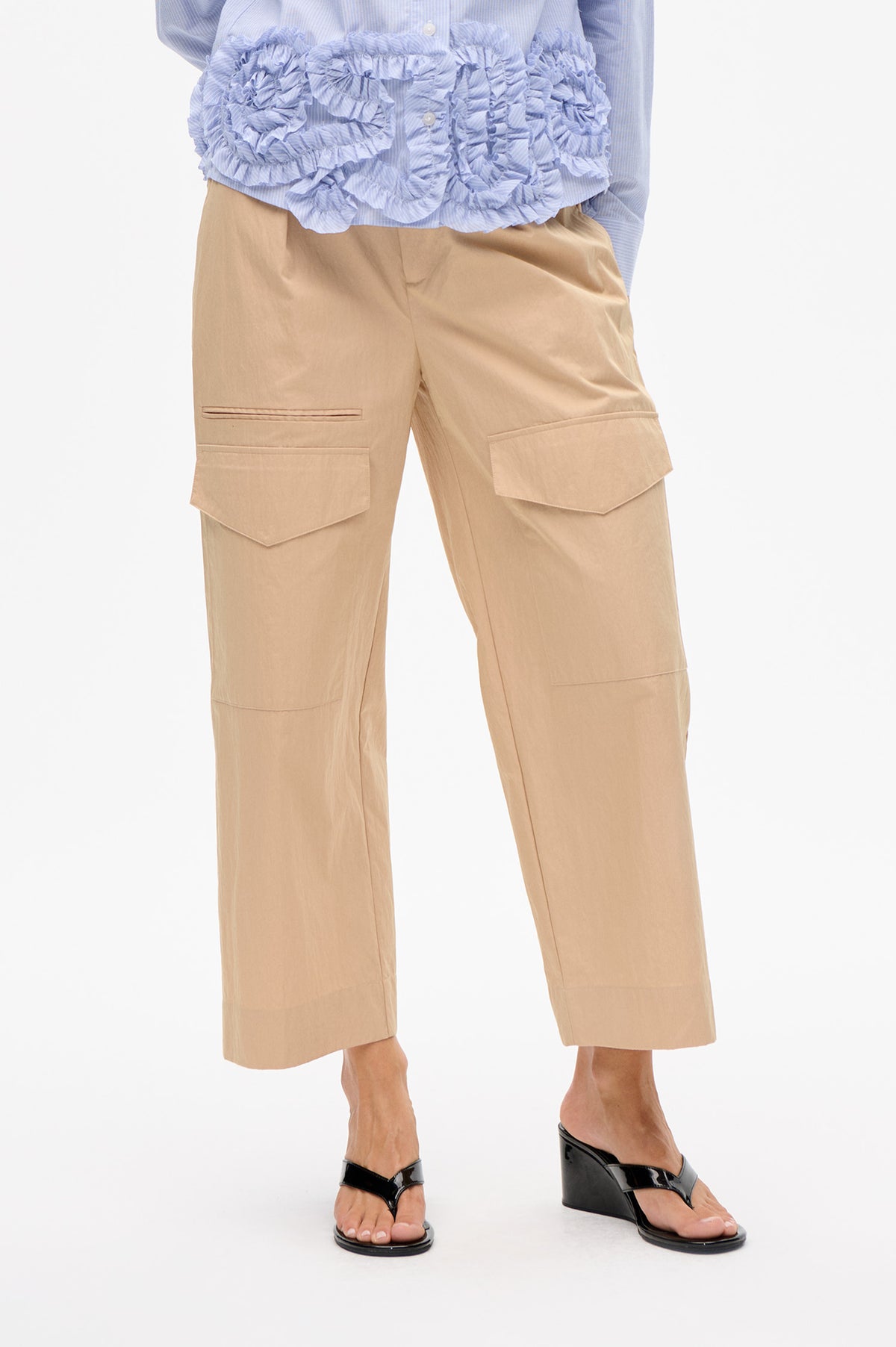 Detailfoto van een beige, wijdvallende broek in kort model met grote klepzakken in cargostijl, gecombineerd met een blauw-wit fijn gestreepte blouse met ruches.