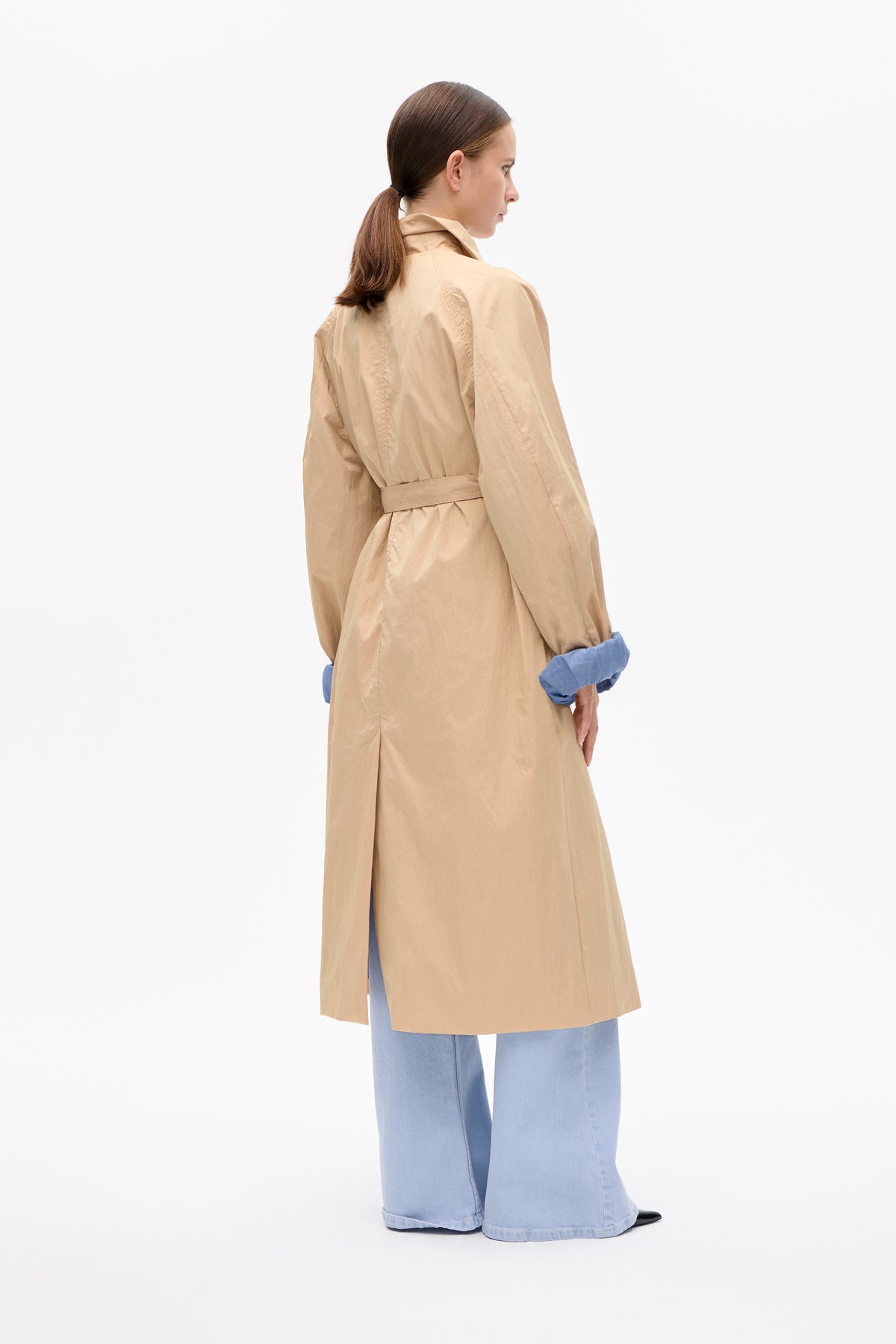 Back view of a model wearing a sand-coloured belted trench coat with turned-up blue cuffs and light blue wide-leg trousers.