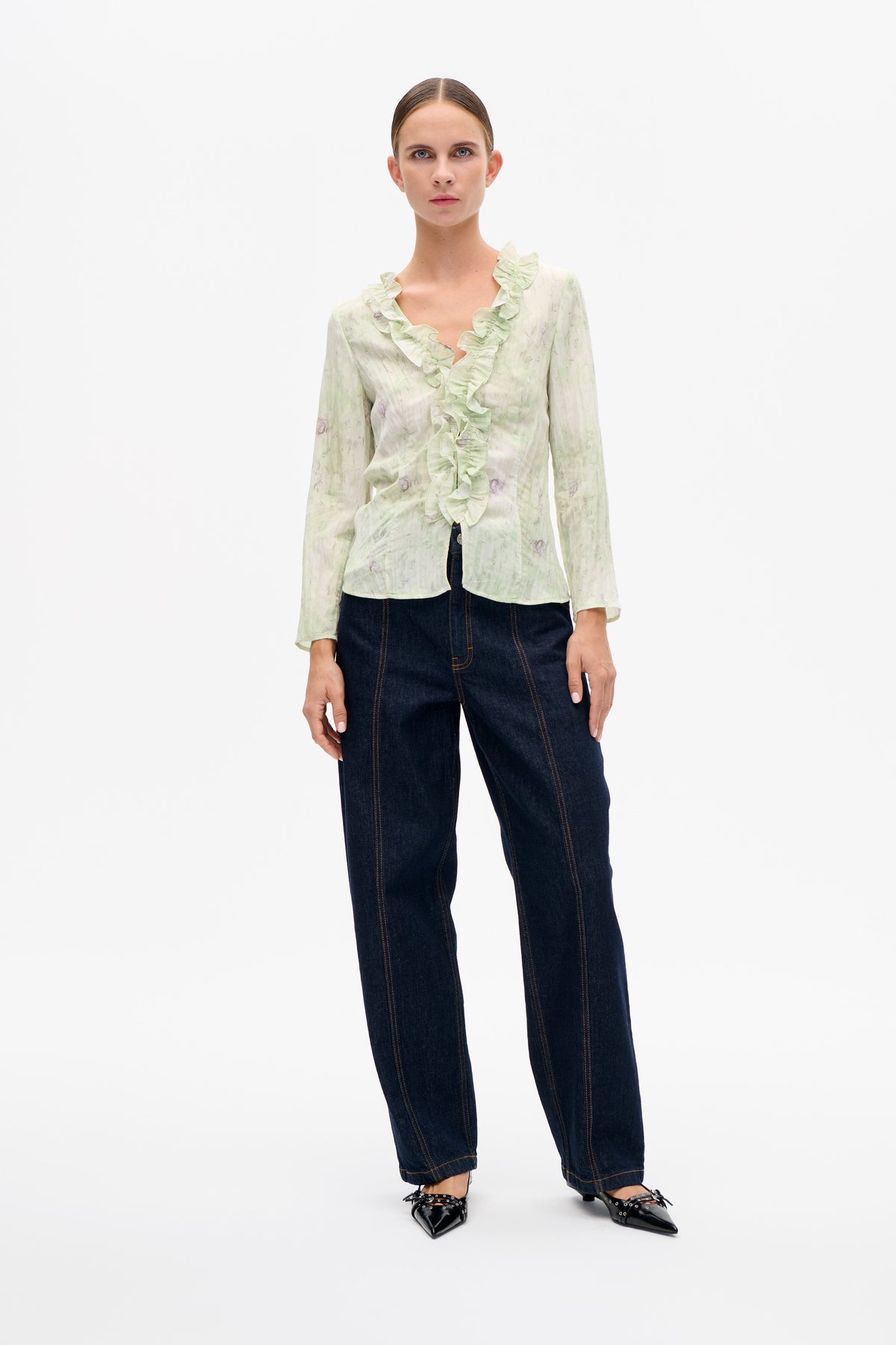 Model draagt een lichtgroene blouse met een subtiel dessin, lange mouwen en ruches langs de V-hals, gecombineerd met donkere jeans.