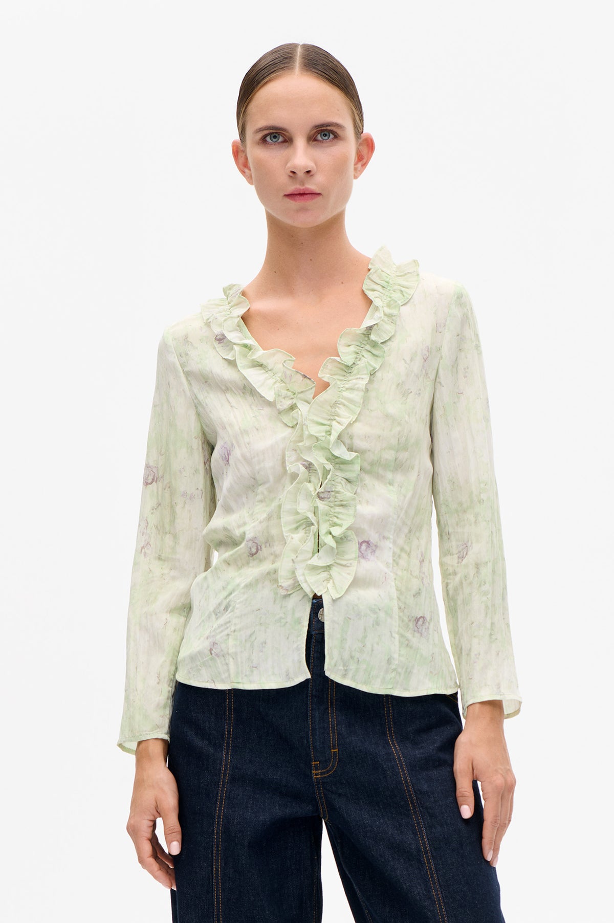 Front view of a pale green and cream printed blouse with long sleeves and a deep V-neck edged in ruffles.