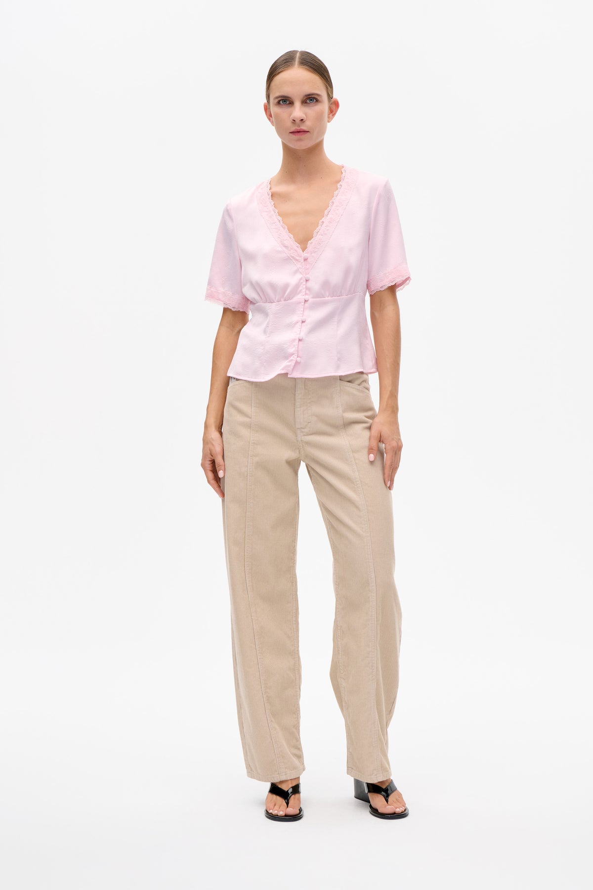 Front view of a model wearing a pale pink short-sleeve blouse with a lace-trimmed V-neck and covered buttons, paired with beige trousers.