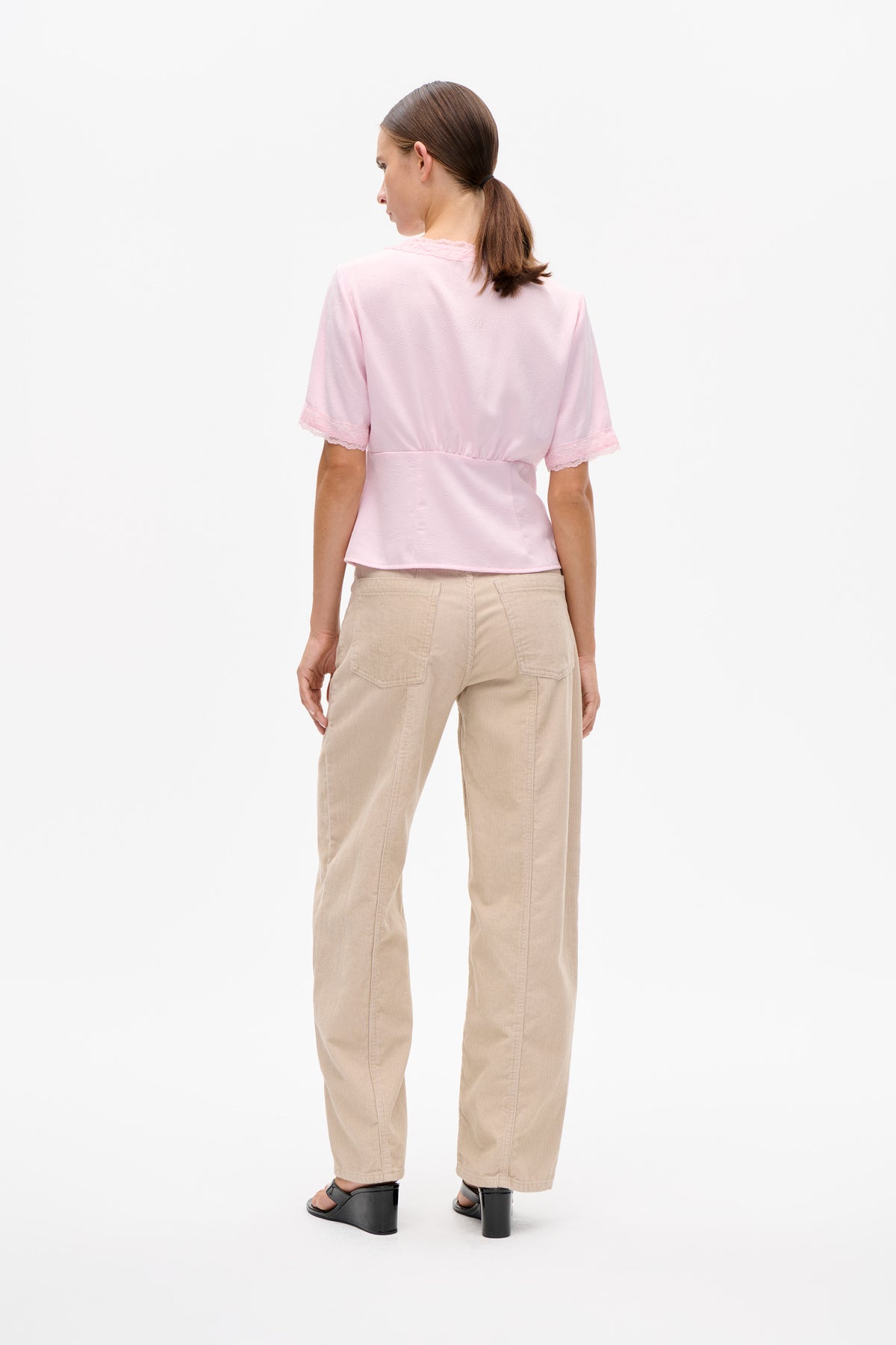 Back view of a model wearing a pale pink short-sleeved blouse with a subtle textured pattern and lace trim at the collar and cuffs, with beige trousers.