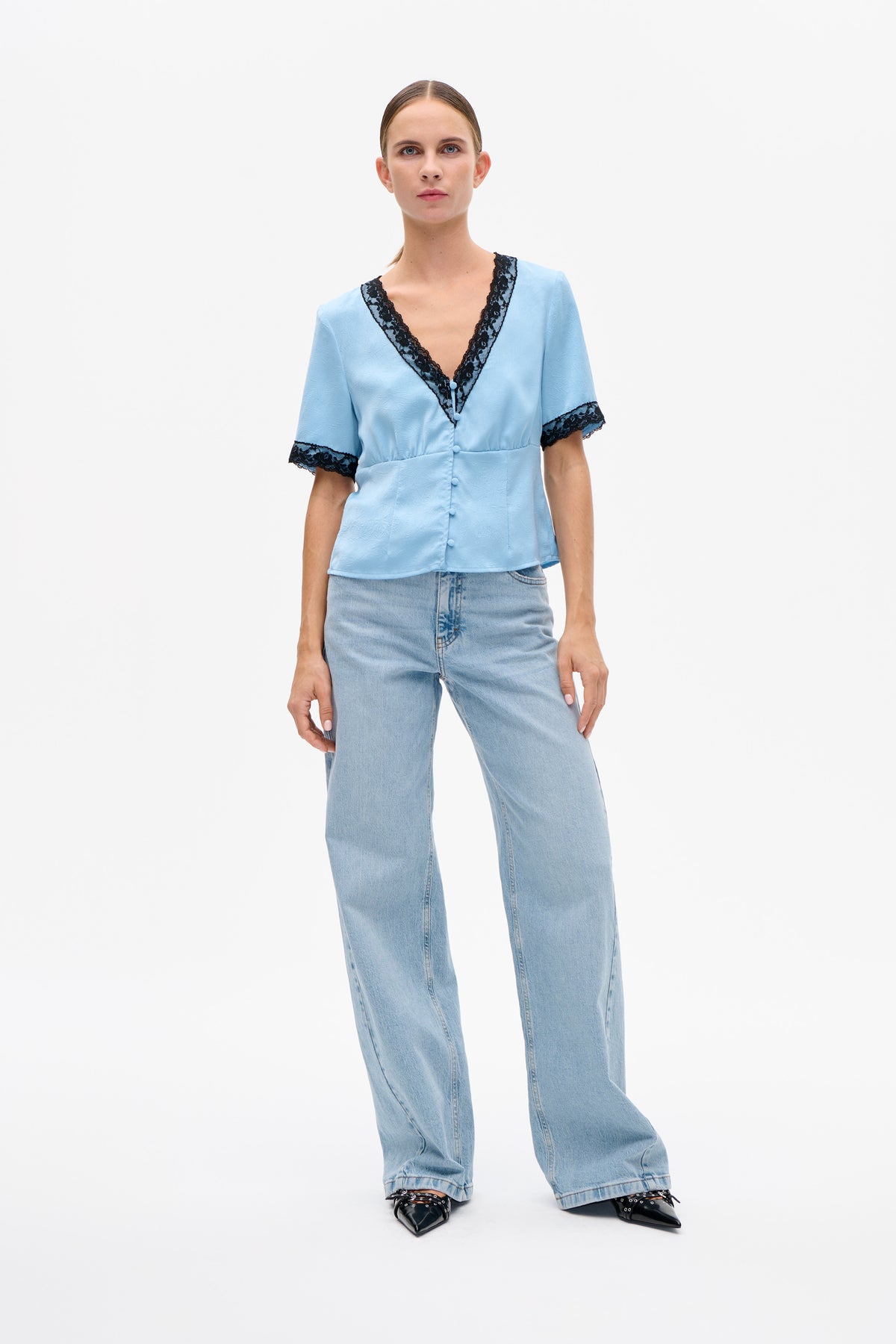 Vooraanzicht van een lichtblauwe blouse met korte mouwen, zwarte kantenbies, een diepe V-hals en stofbeklede knopen, gecombineerd met wijde jeans.