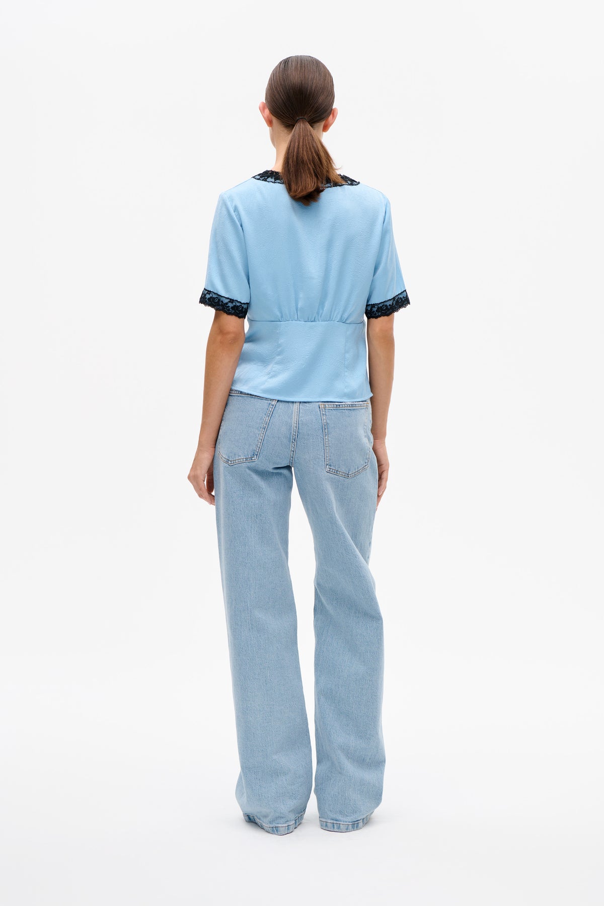Achteraanzicht van een lichtblauwe blouse met korte mouwen en zwarte kantafwerking bij kraag en manchetten, gedragen met wijdvallende lichtgewassen jeans.