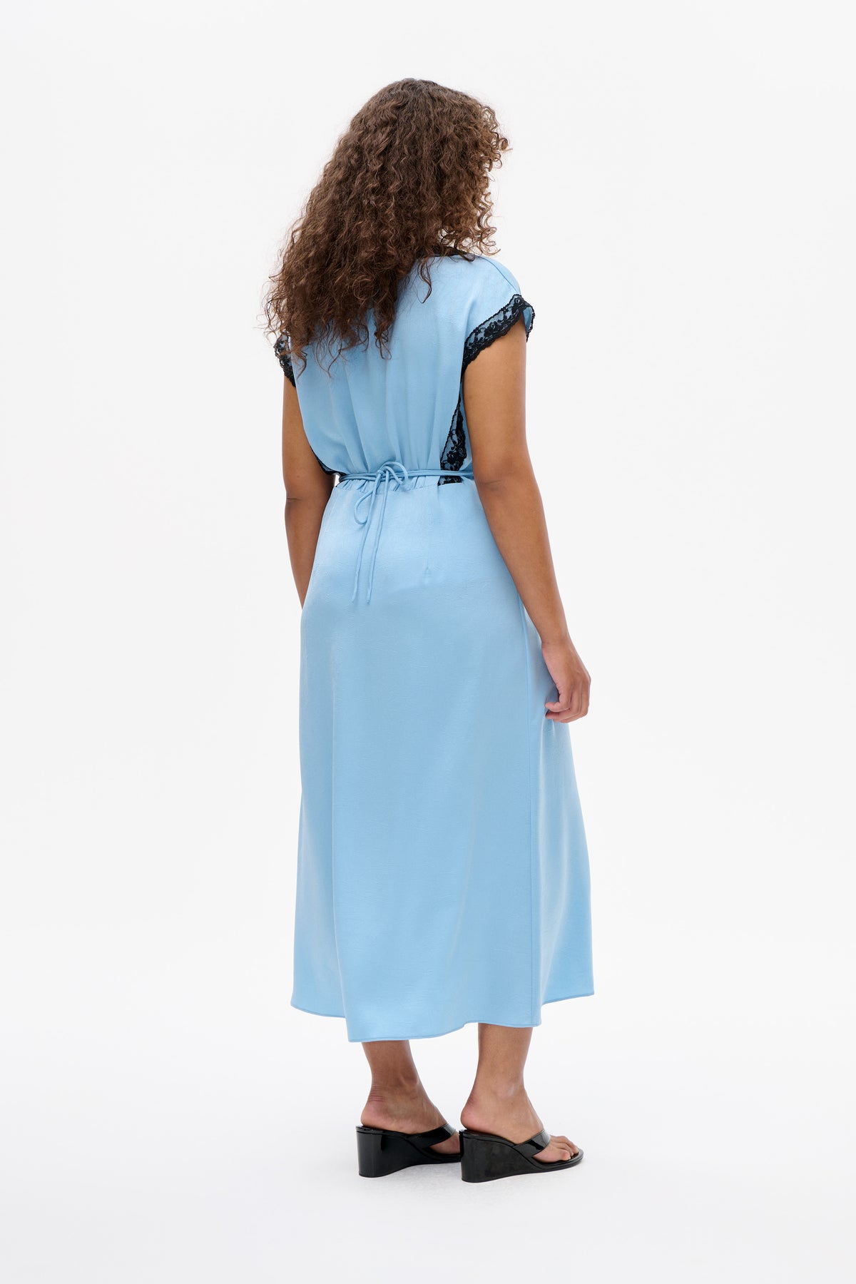Back view of a light blue midi dress with black lace sleeve and side trim and a tie belt at the waist.