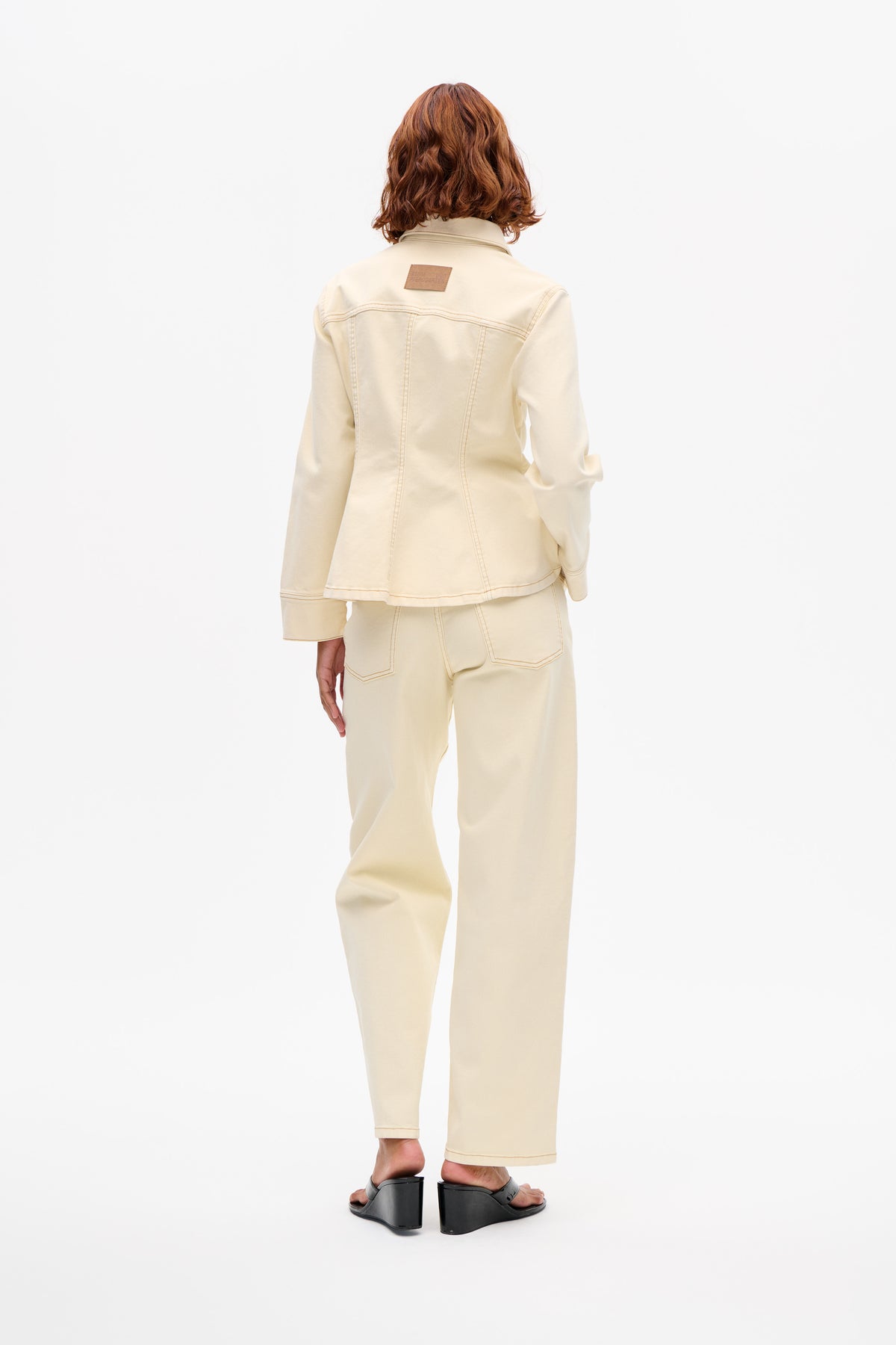 Vue arrière d'un mannequin portant une veste crème à col, avec surpiqûres contrastées courbes et un patch marron au dos, portée avec un pantalon coordonné.