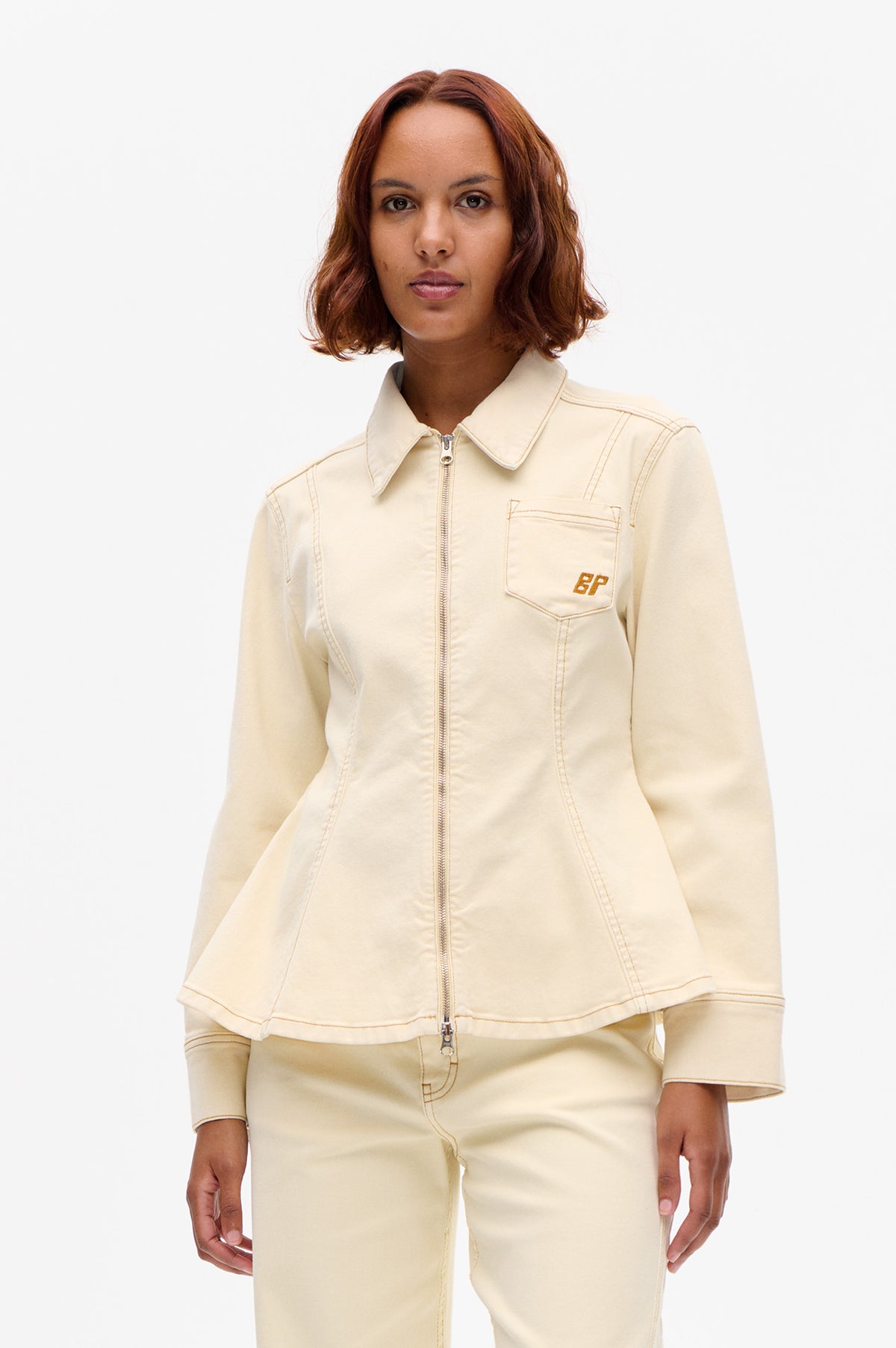 Front view of a cream zip-front jacket with pointed collar, contrast stitching and a chest pocket bearing a BP monogram, worn with matching trousers.
