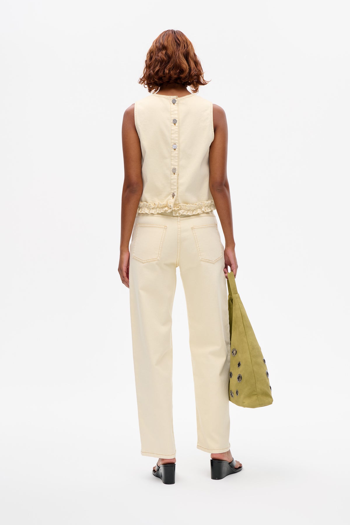 Rear view of a model in a cream sleeveless top with back buttons and a ruffled hem, paired with wide-leg trousers.