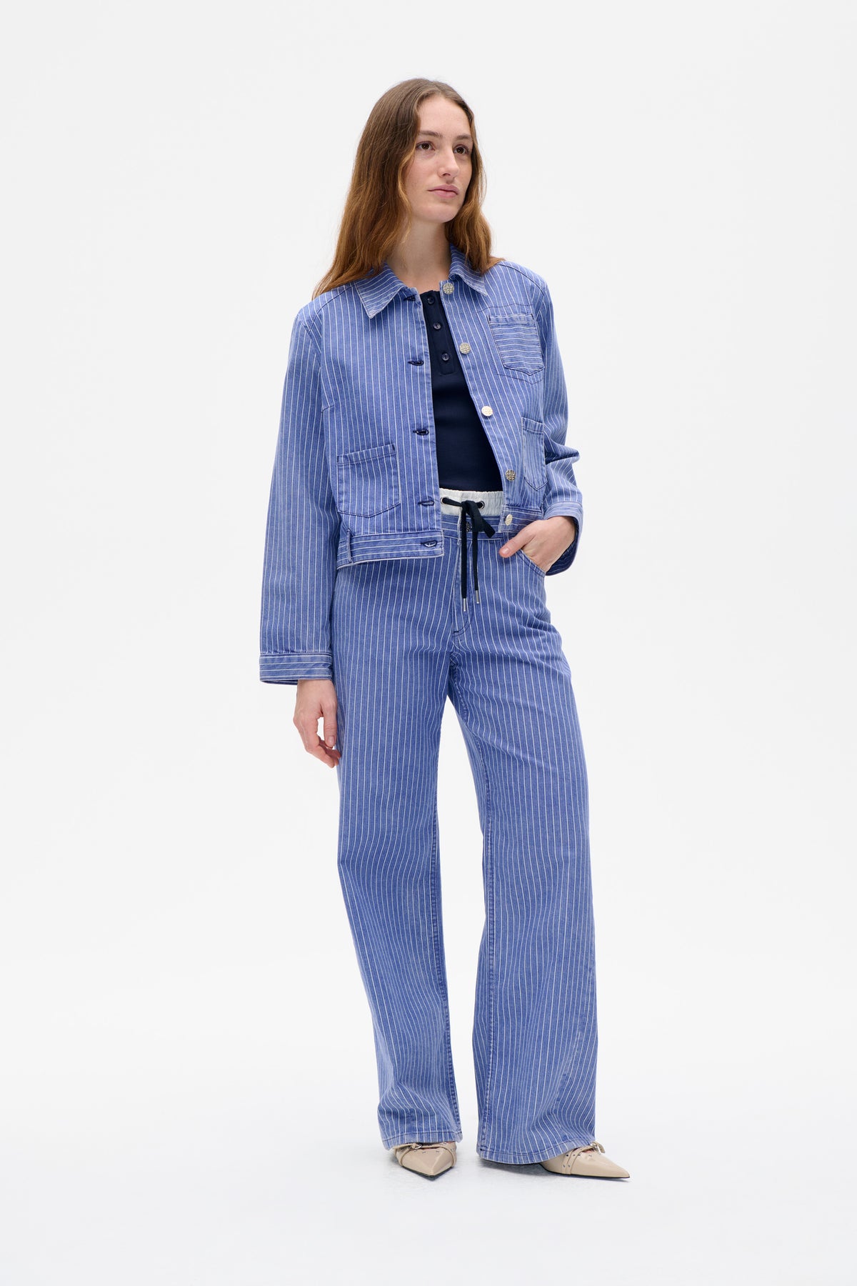 Model wearing a blue-and-white pinstriped utility jacket with matching wide-leg trousers and a navy top, shown from the front.