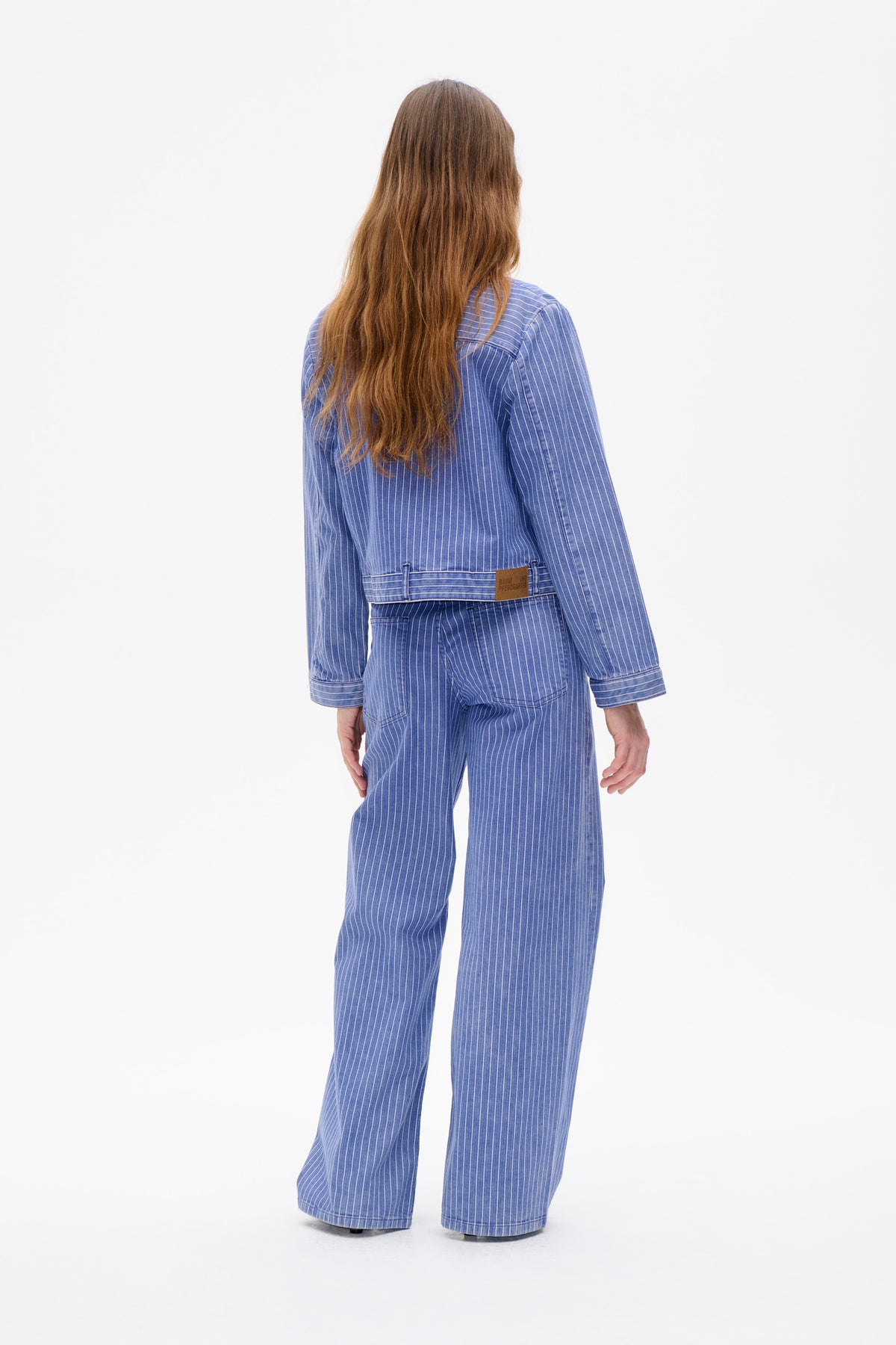 Rear view of a model in a blue-and-white pinstripe jacket and wide-leg trousers, with a back waist tab and leather logo patch.