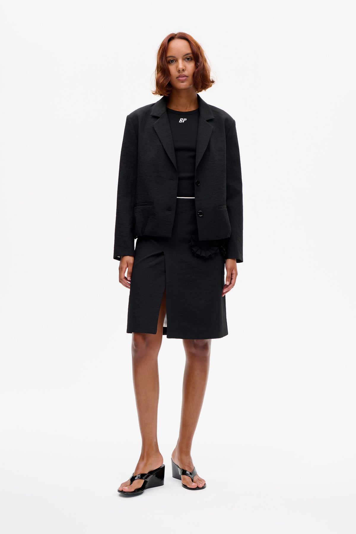 Front-facing studio styling of a charcoal blazer over a black ribbed logo top and knee-length skirt with a ruffled waist detail, paired with glossy black wedge sandals.