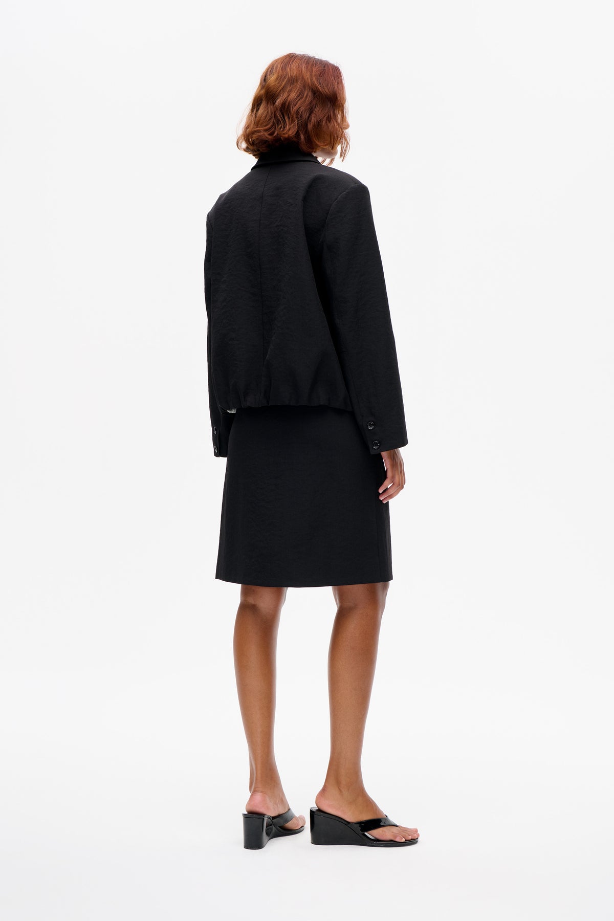Model turned away in a black tailored blazer with buttoned cuffs and a black knee-length skirt, shown from the back against white.