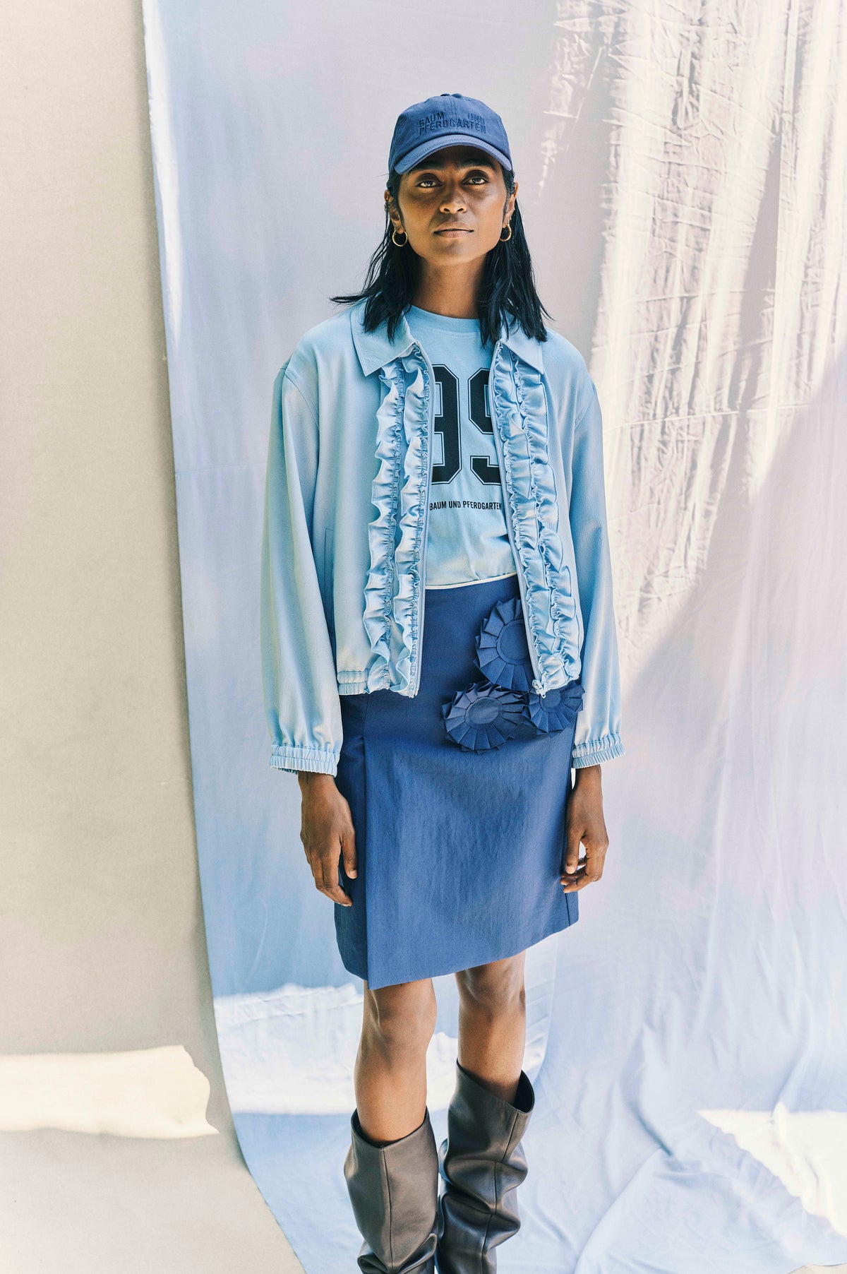 A model wears a blue Baum und Pferdgarten cap, pale blue ruffled jacket, graphic T-shirt, blue skirt and tall black boots.