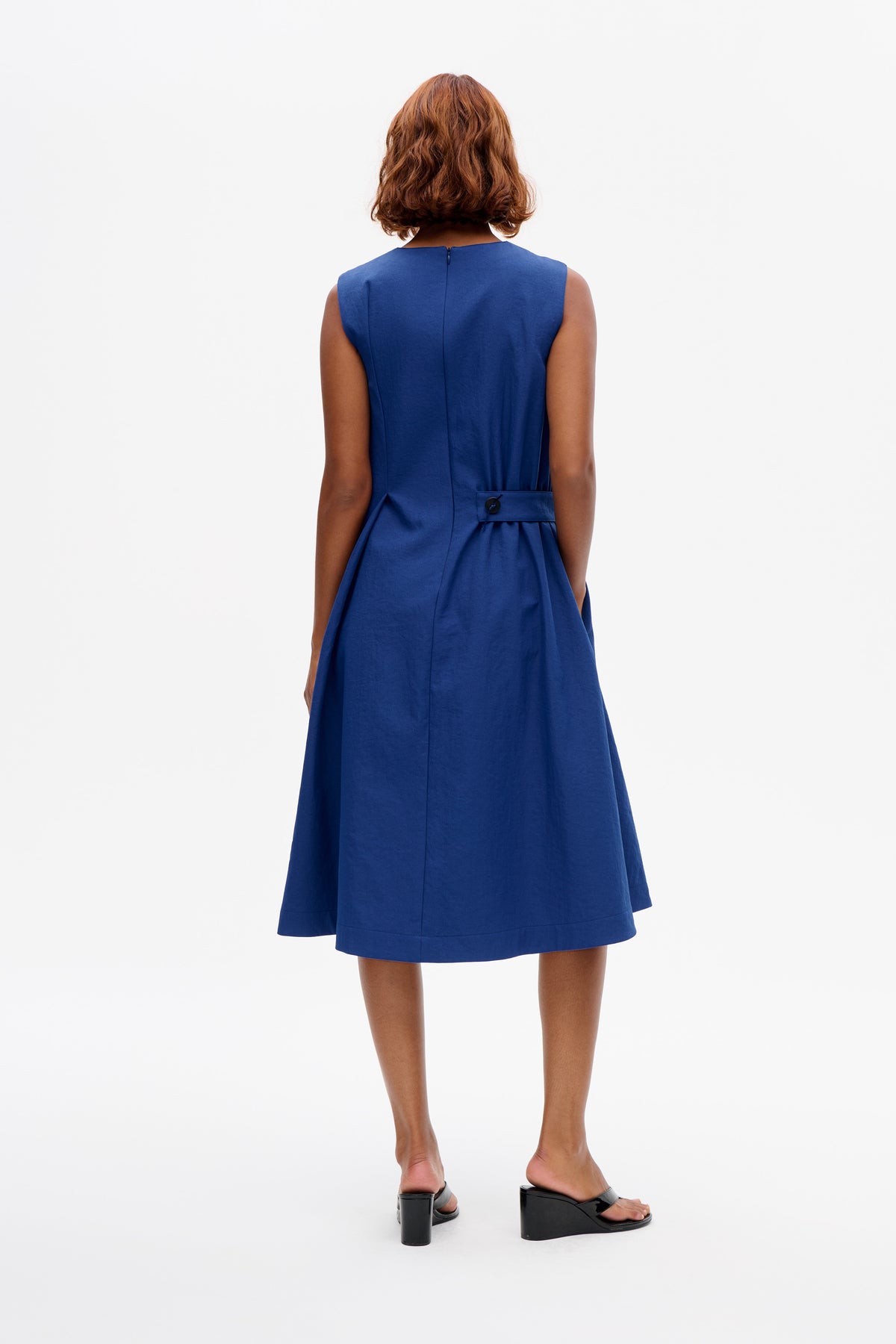 Back view of a model wearing a cobalt blue sleeveless A-line dress with a centre-back zip and buttoned waist tab.