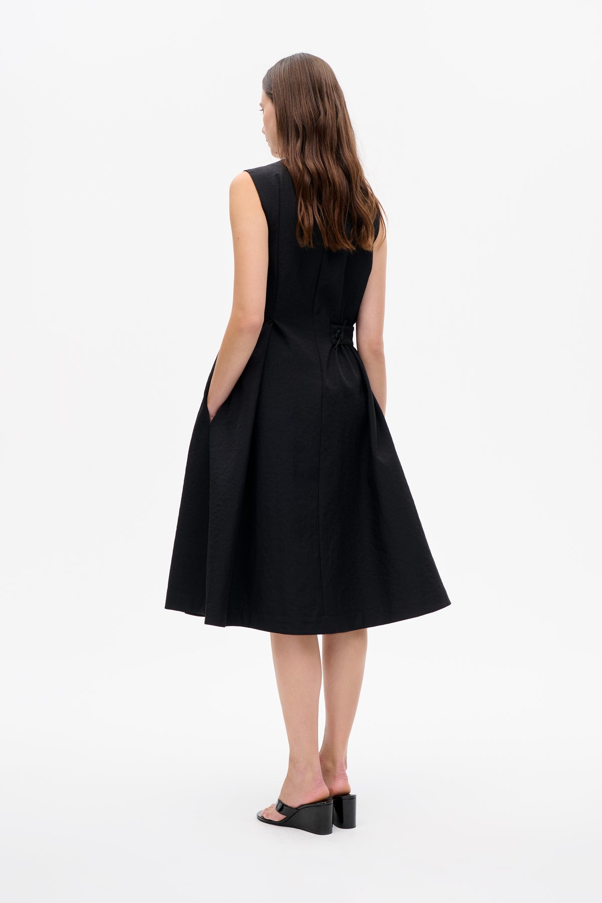 Back view of a model wearing a black sleeveless dress with a fitted waist, button detail and flared knee-length skirt.