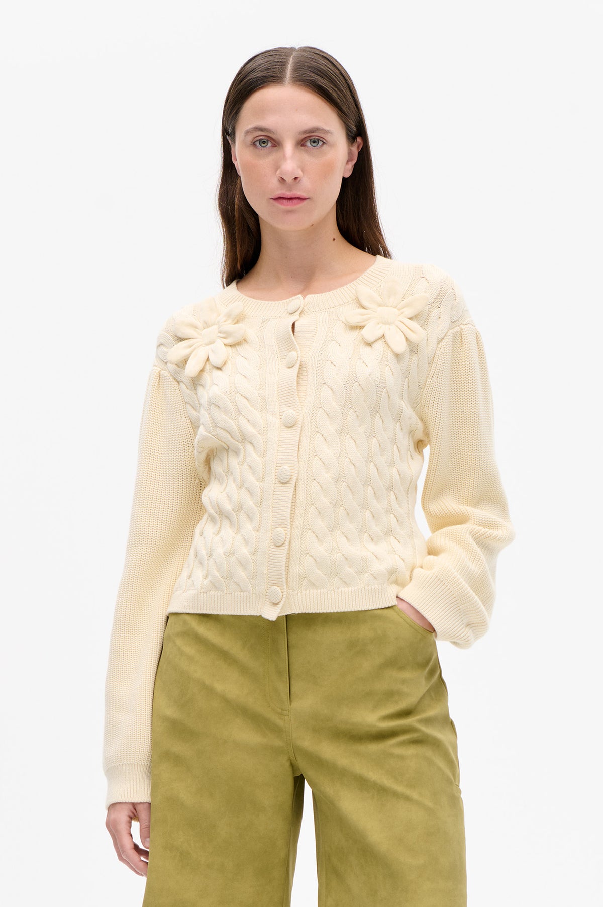 Front view of a cream cable-knit cardigan with floral appliqués and covered buttons, styled with olive-green trousers.