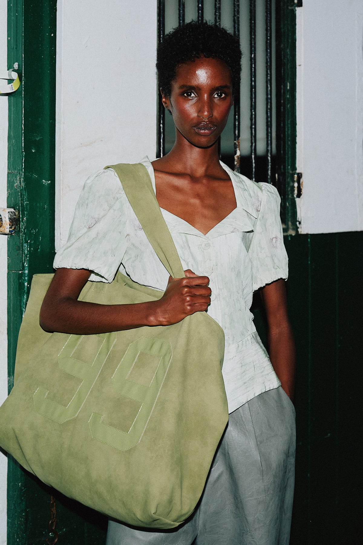 Campaign portrait of a model in a pale floral short-sleeved blouse, carrying a large sage-green tote with stitched lettering.
