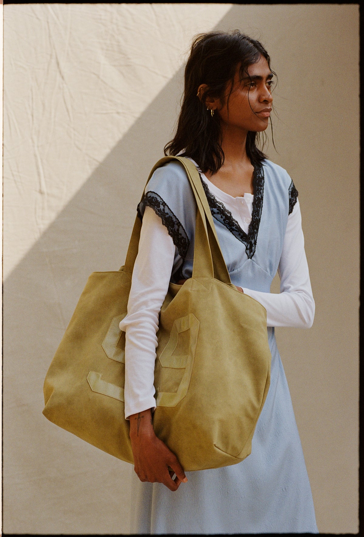 Model in a pale blue sleeveless dress with black lace trim over a white long-sleeved top, carrying a large mustard tote.