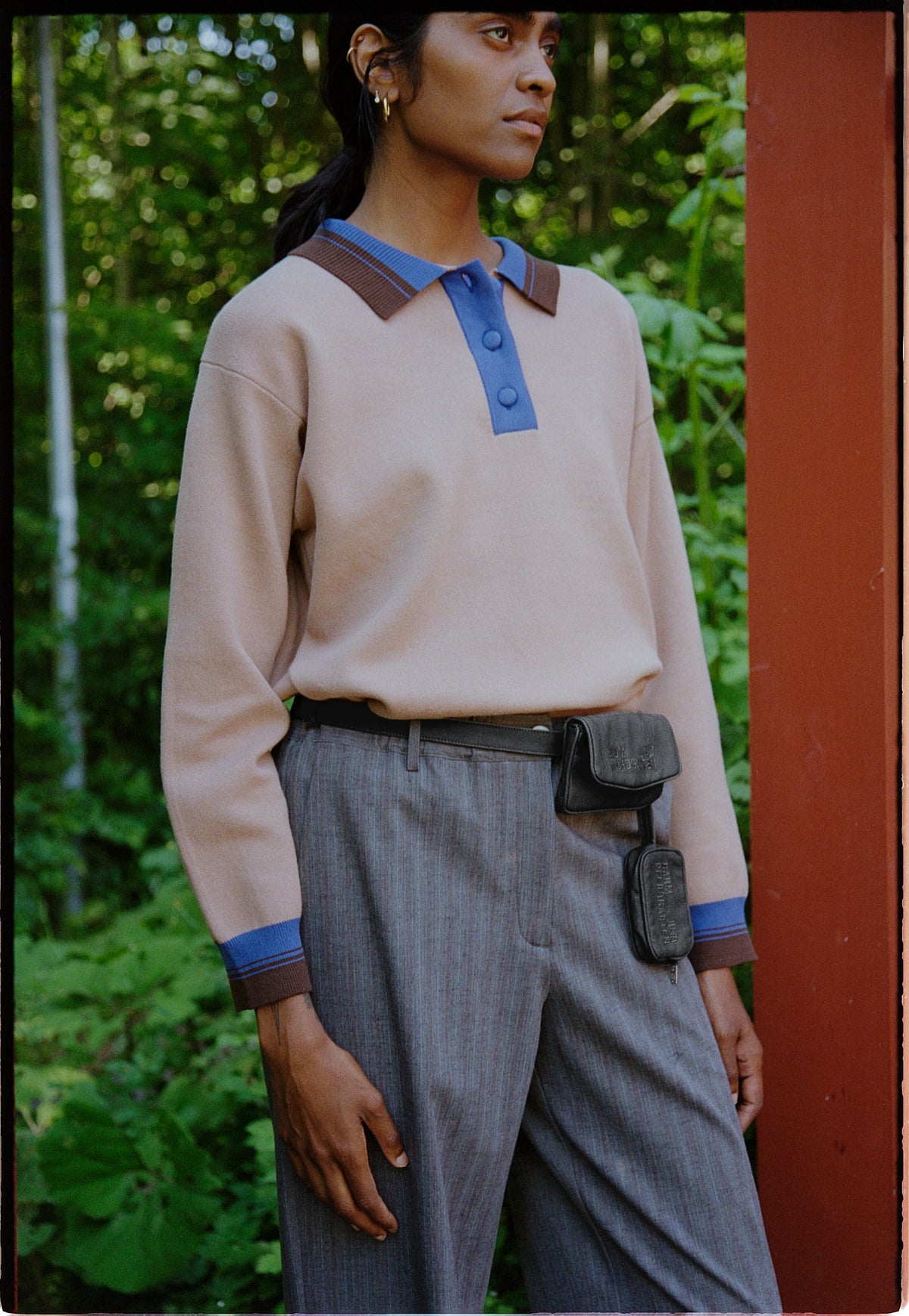 Model wearing a cream long-sleeved top with a blue button placket and striped collar, paired with grey trousers and a black belt bag.