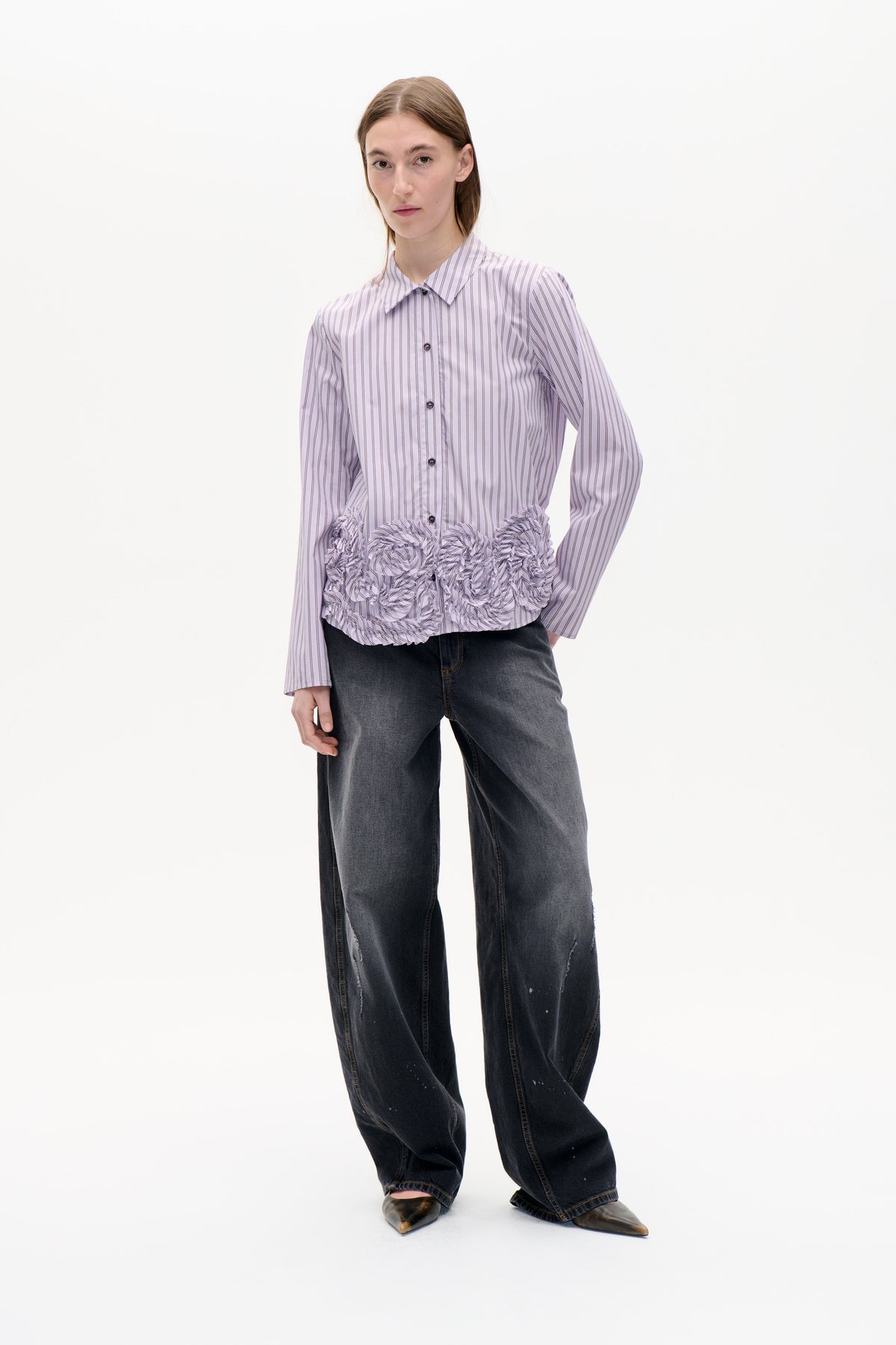 Model wearing a pale lavender button-front shirt with fine dark vertical stripes and gathered ruffle detailing across the lower front.