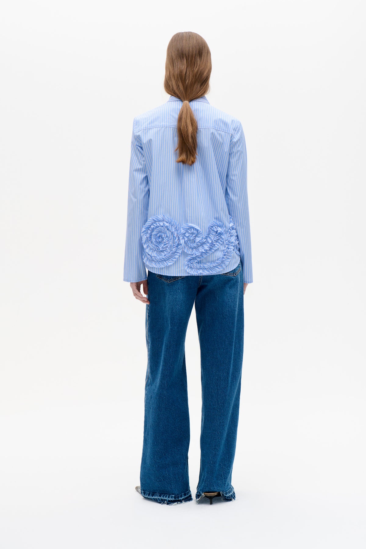 Achteraanzicht van een blauw-wit gestreepte blouse met lange mouwen en oversized, geplooide ruches op de onderrug, gecombineerd met donkere jeans met wijde pijpen.