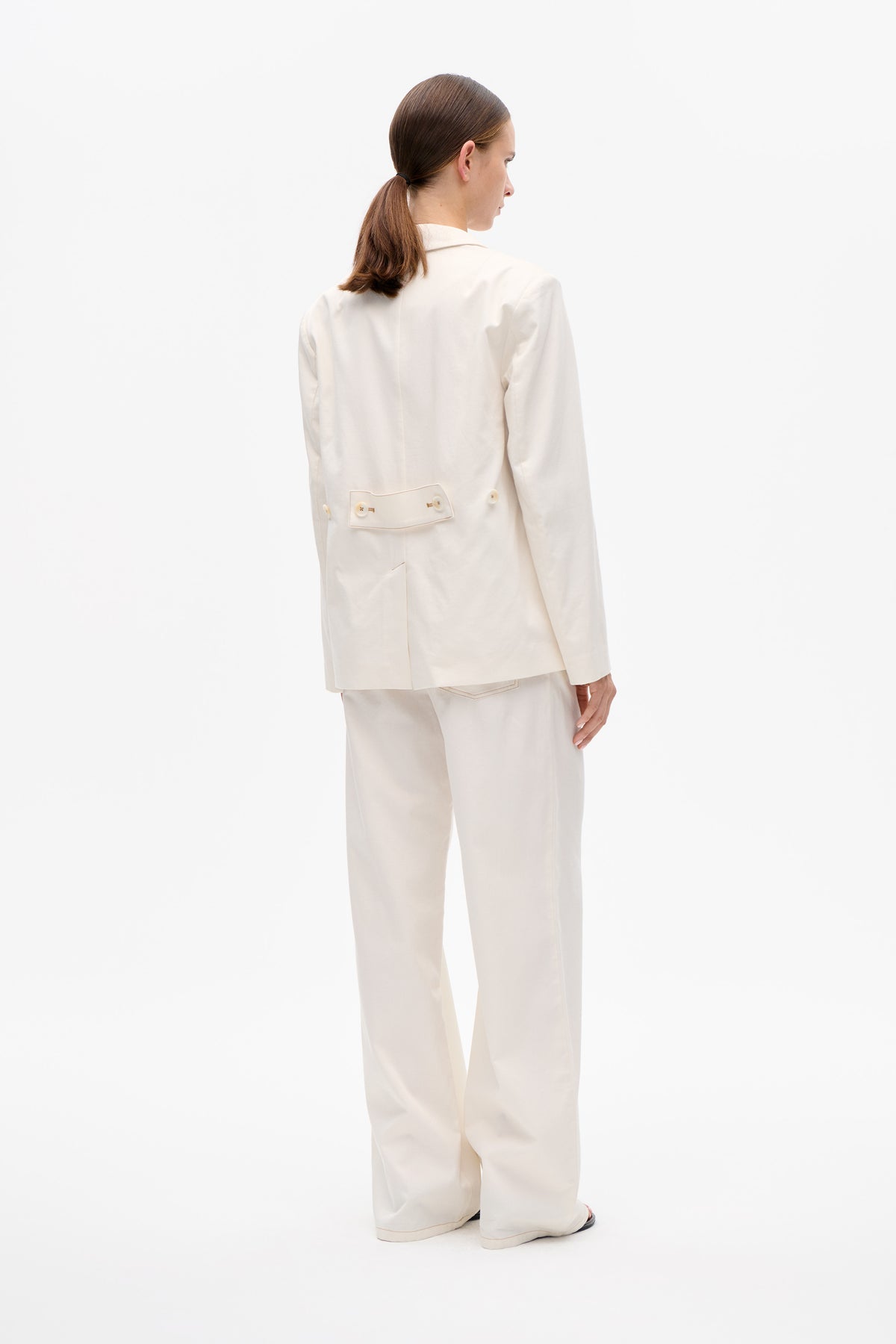 Back view of a model wearing an ivory blazer with a buttoned waist tab and matching wide-leg trousers.