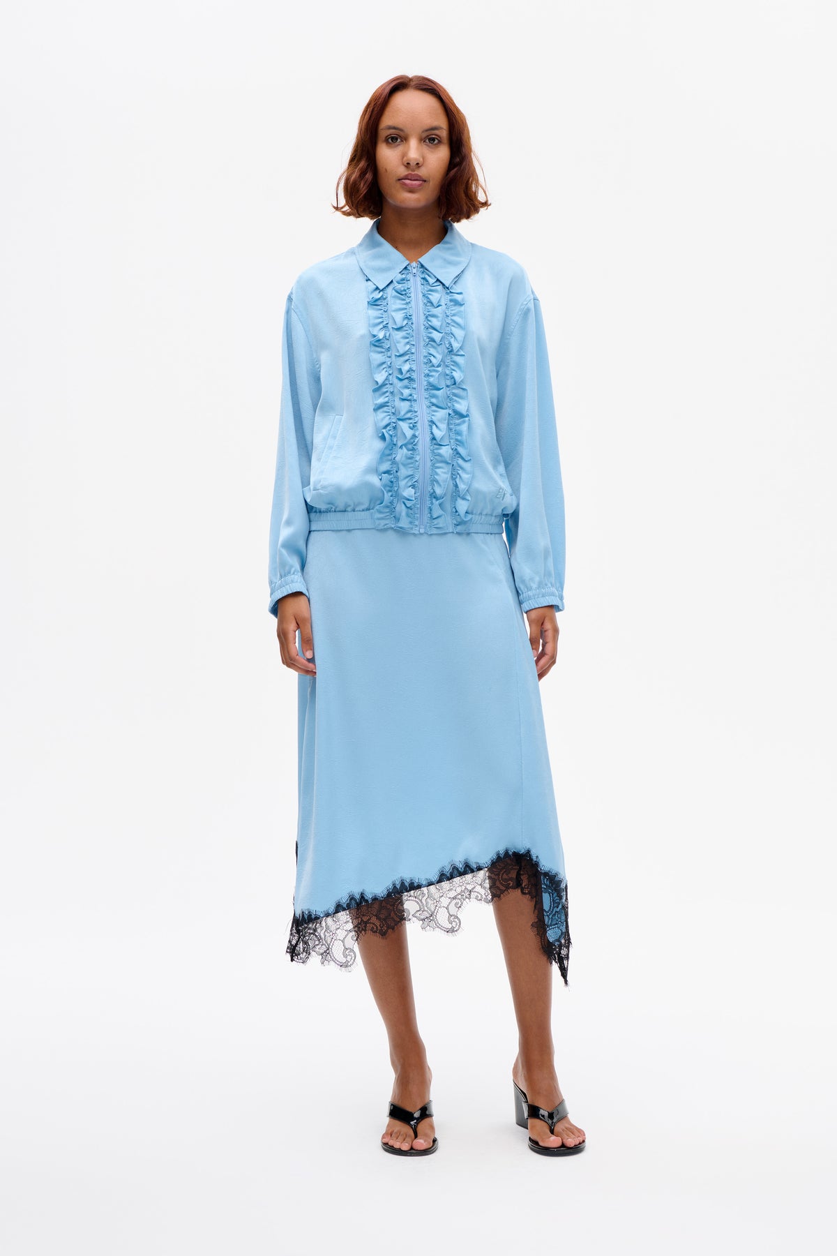 Front-facing model wearing a light blue ruffle-trim zip jacket over a matching dress with a black lace hem.