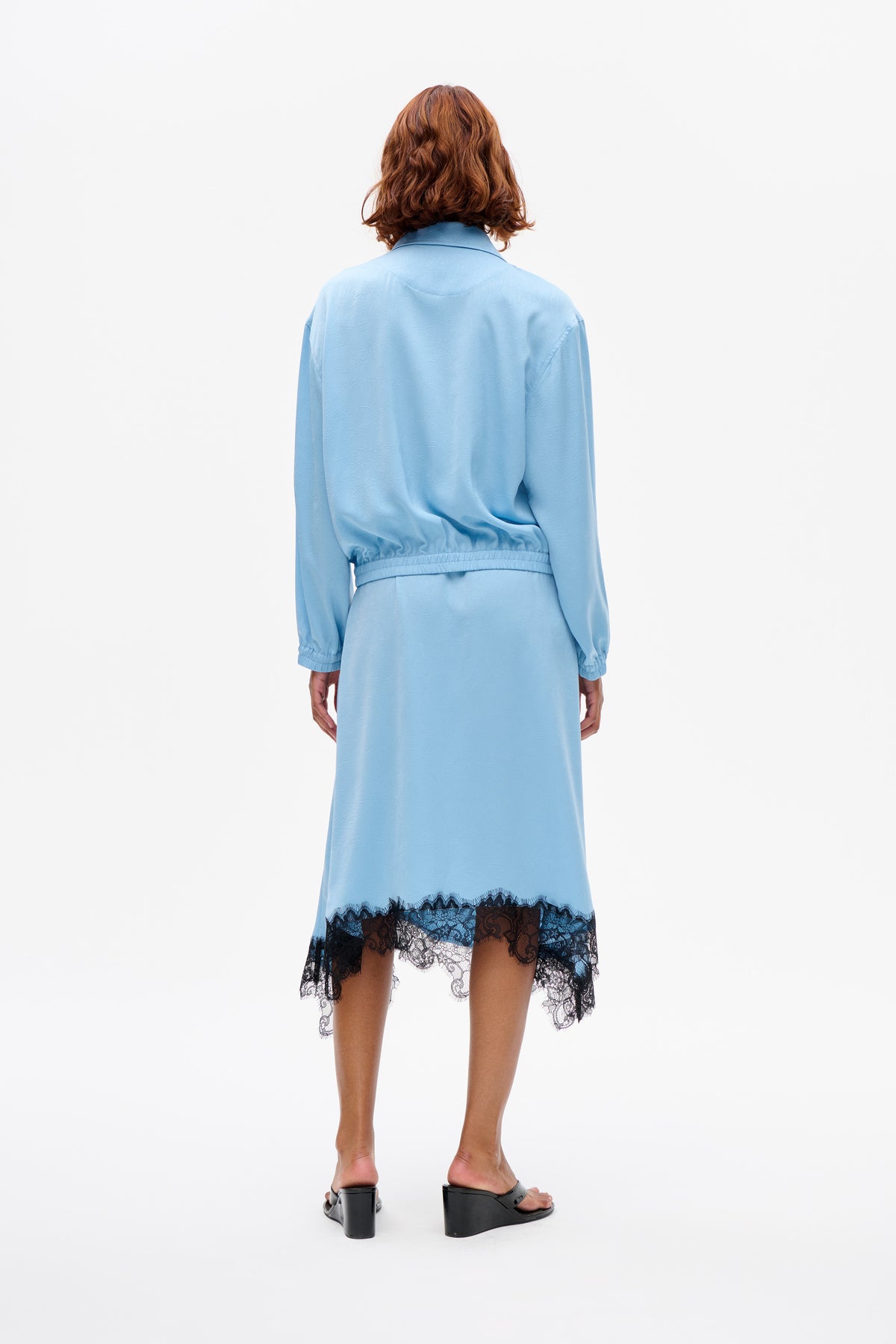 Back view of a model in a light blue textured jacket and matching skirt with a black lace hem.