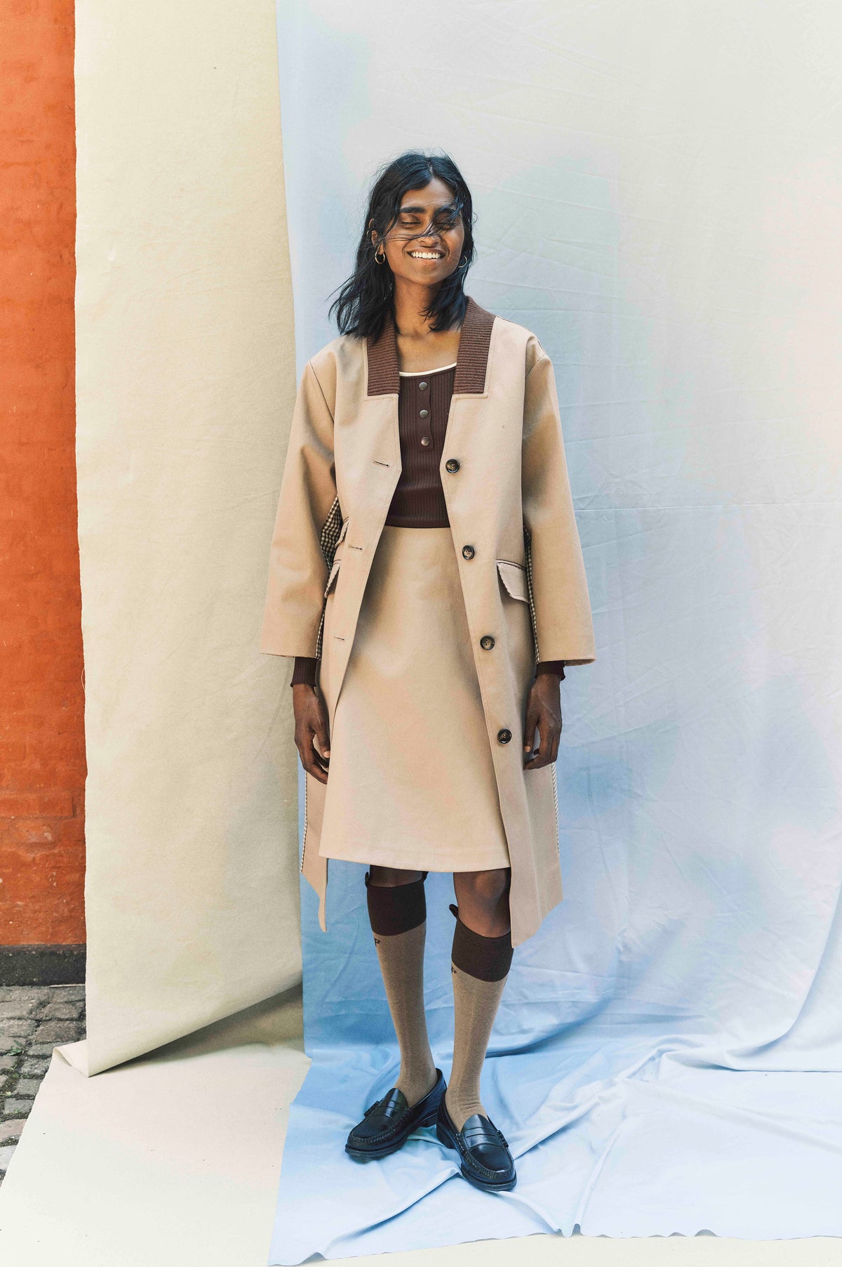 Campaign portrait of a smiling model in a long beige button-front coat with a wide brown ribbed collar, brown top, knee socks and black loafers.