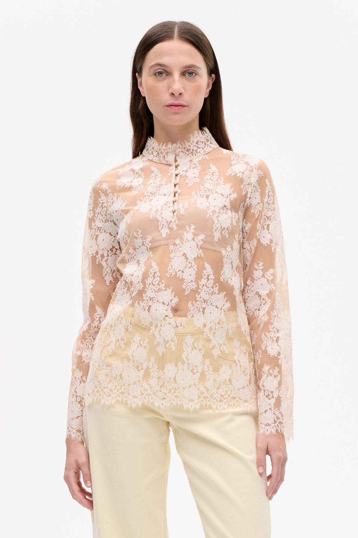 Front view of a sheer blush blouse with white floral lace, a high lace collar and fabric-covered buttons, paired with cream trousers.