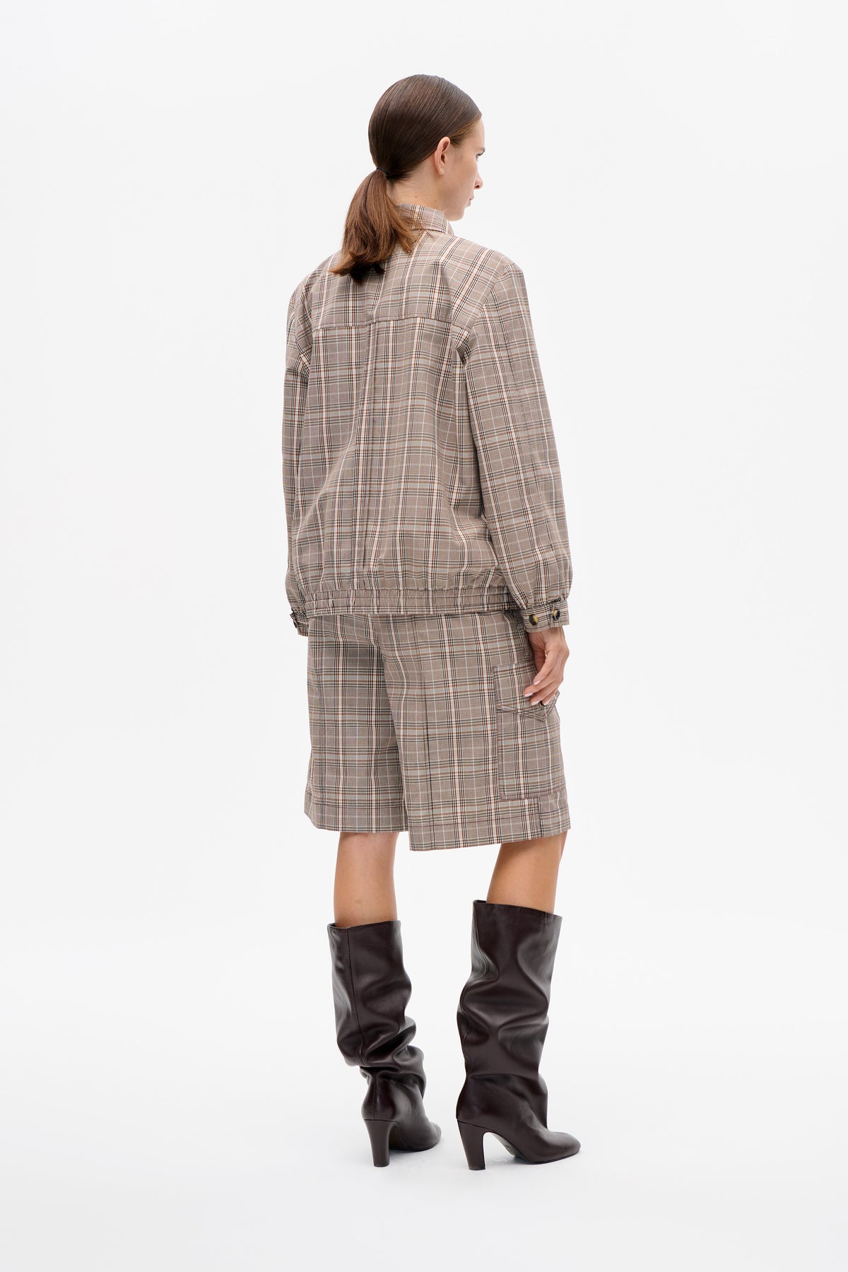 Back view of a model wearing a beige plaid jacket and matching shorts with dark brown knee-high heeled boots.