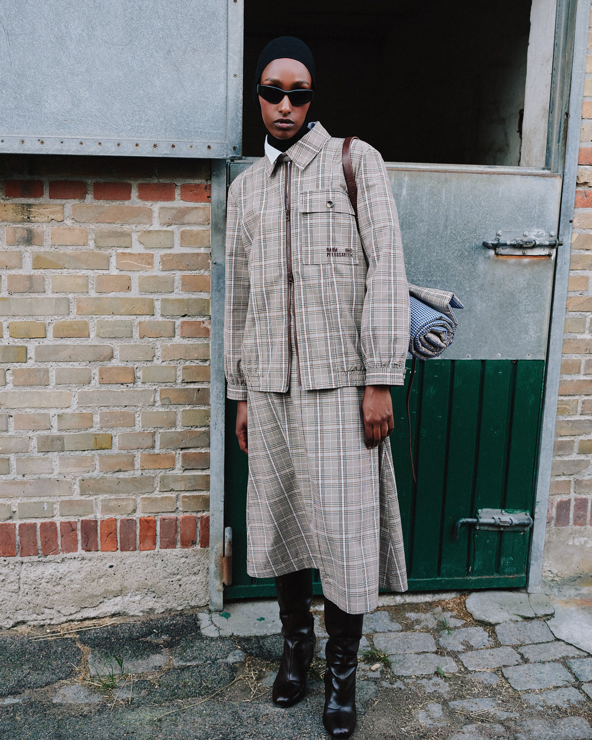 Model wearing a beige plaid button-front jacket and matching long skirt, styled with black boots and sunglasses.